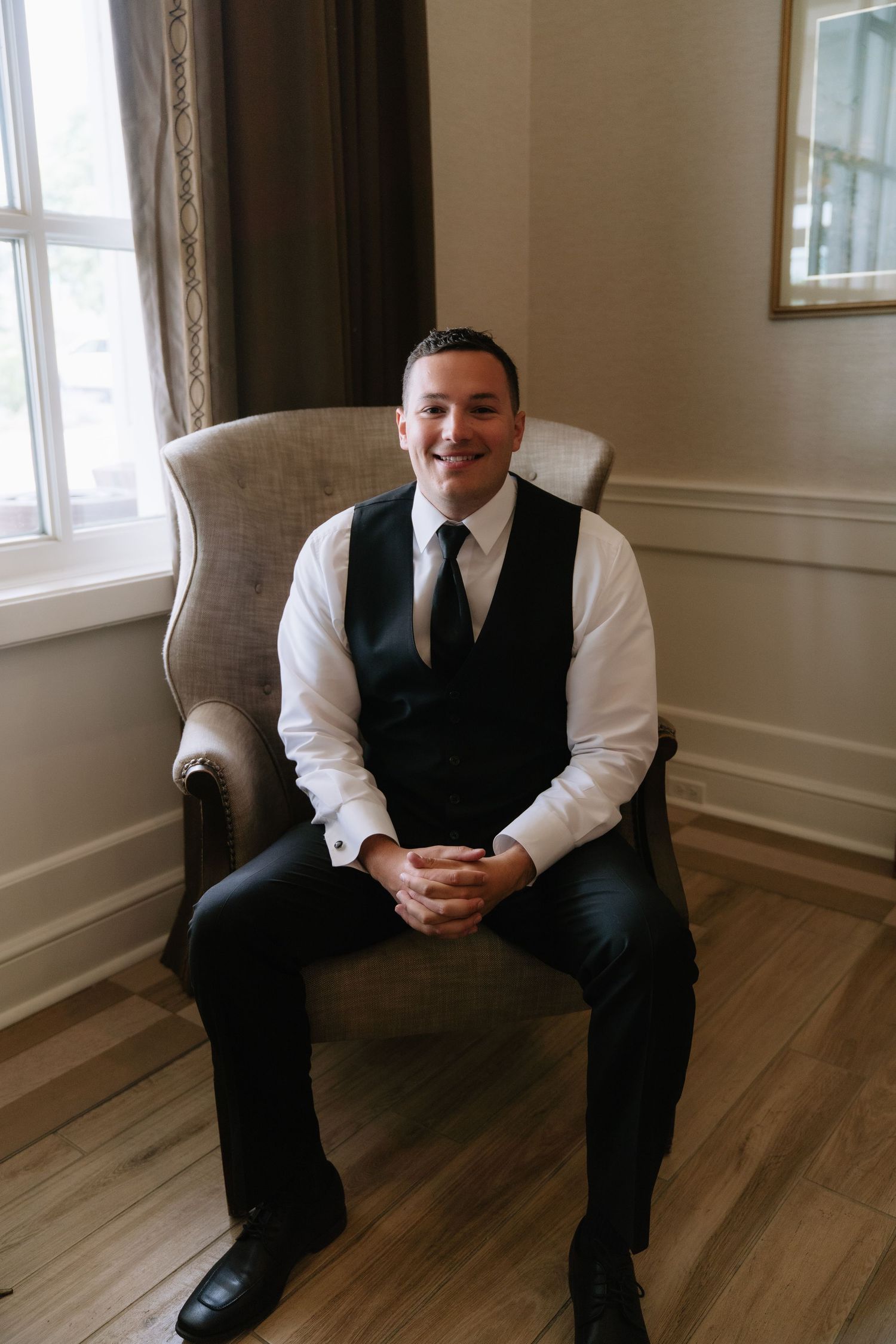 Smiling groom in black vest and tie sits in elegant chair, hands clasped, posing for wedding day portrait.