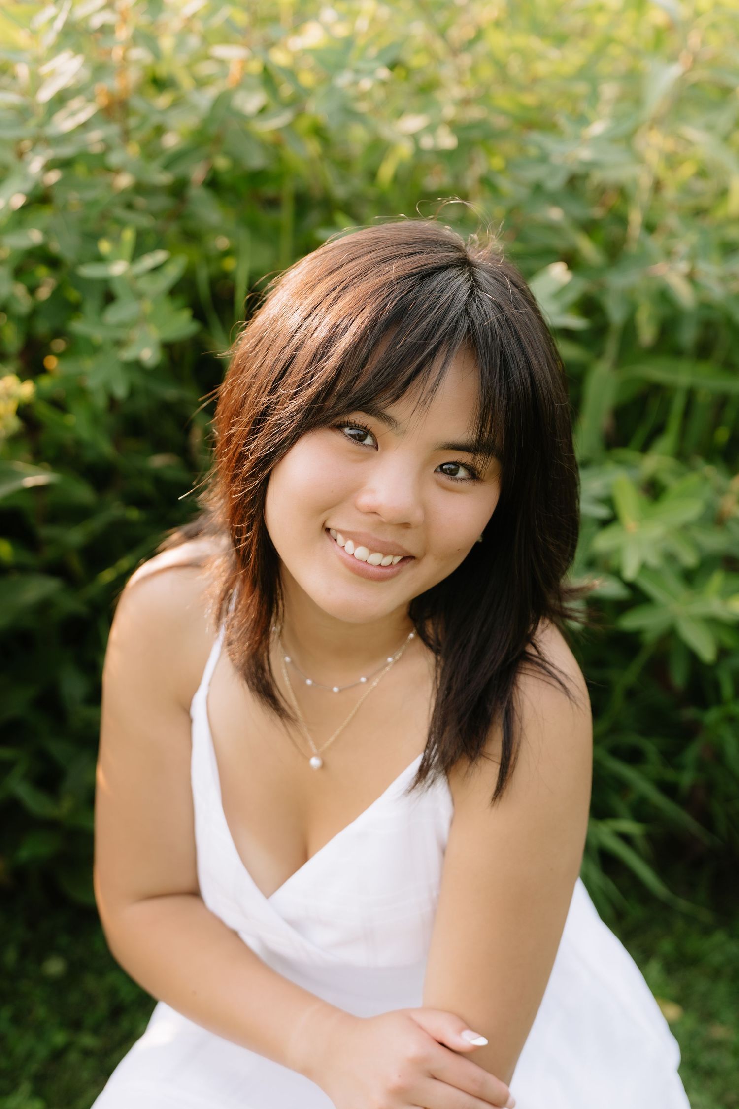 Smiling young woman in white dress with necklace sits outdoors among lush green foliage.