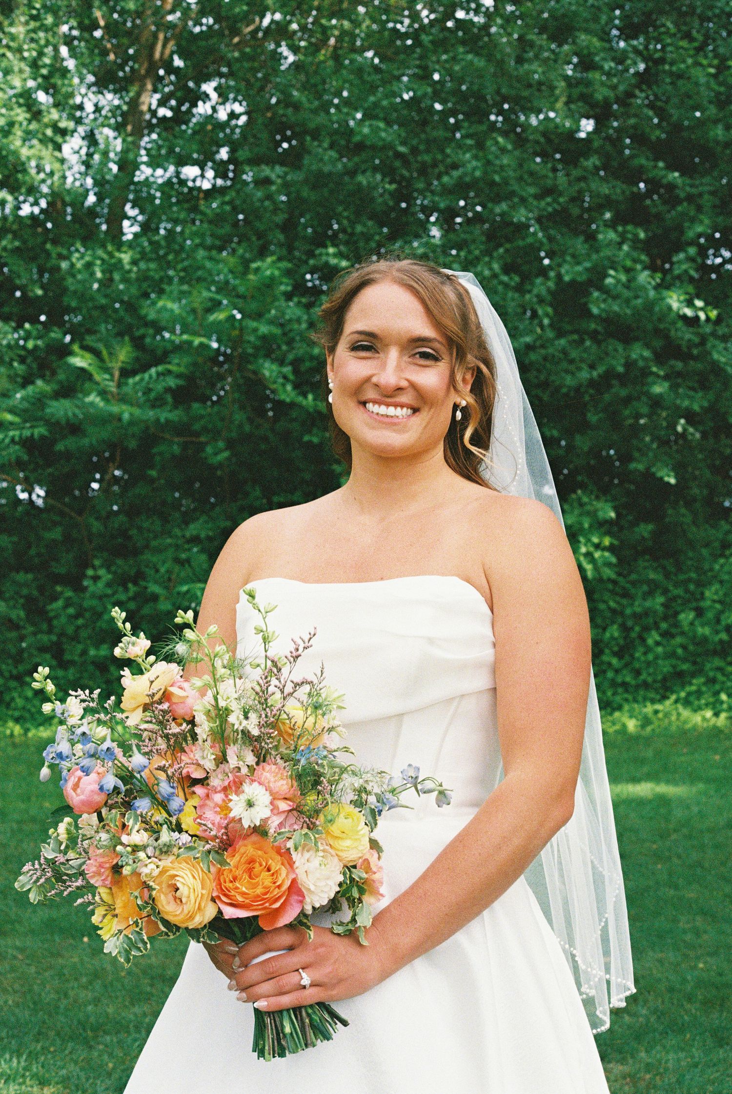 Smiling bride in strapless white gown holding colorful spring bouquet with orange, pink, and blue flowers outdoors.