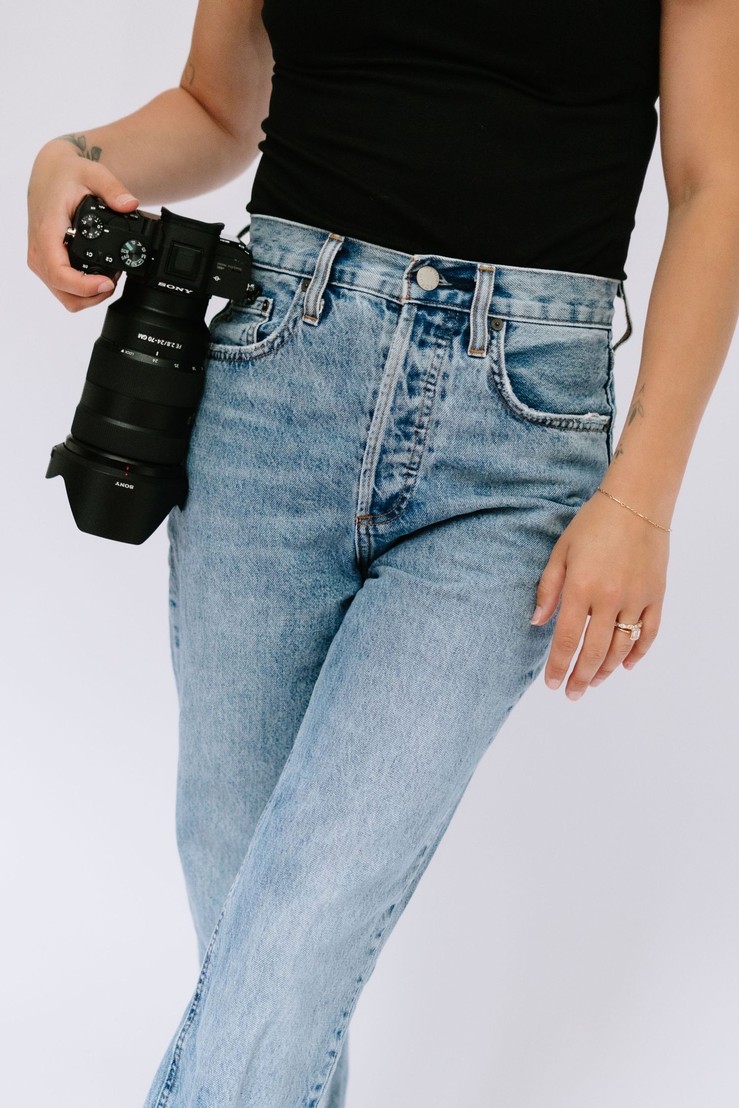 Female photographer in black top and light wash jeans holding a professional DSLR camera.