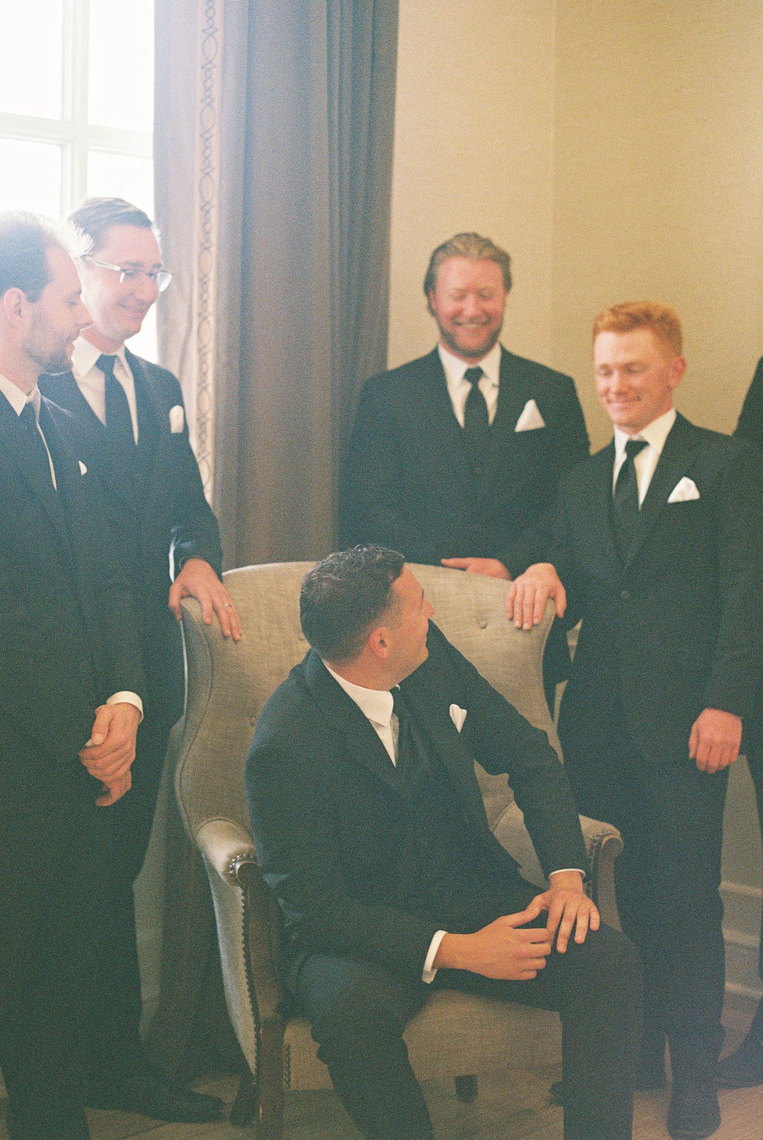 Groomsmen in black suits gather around groom seated in gray chair, sharing a joyful moment before wedding ceremony.