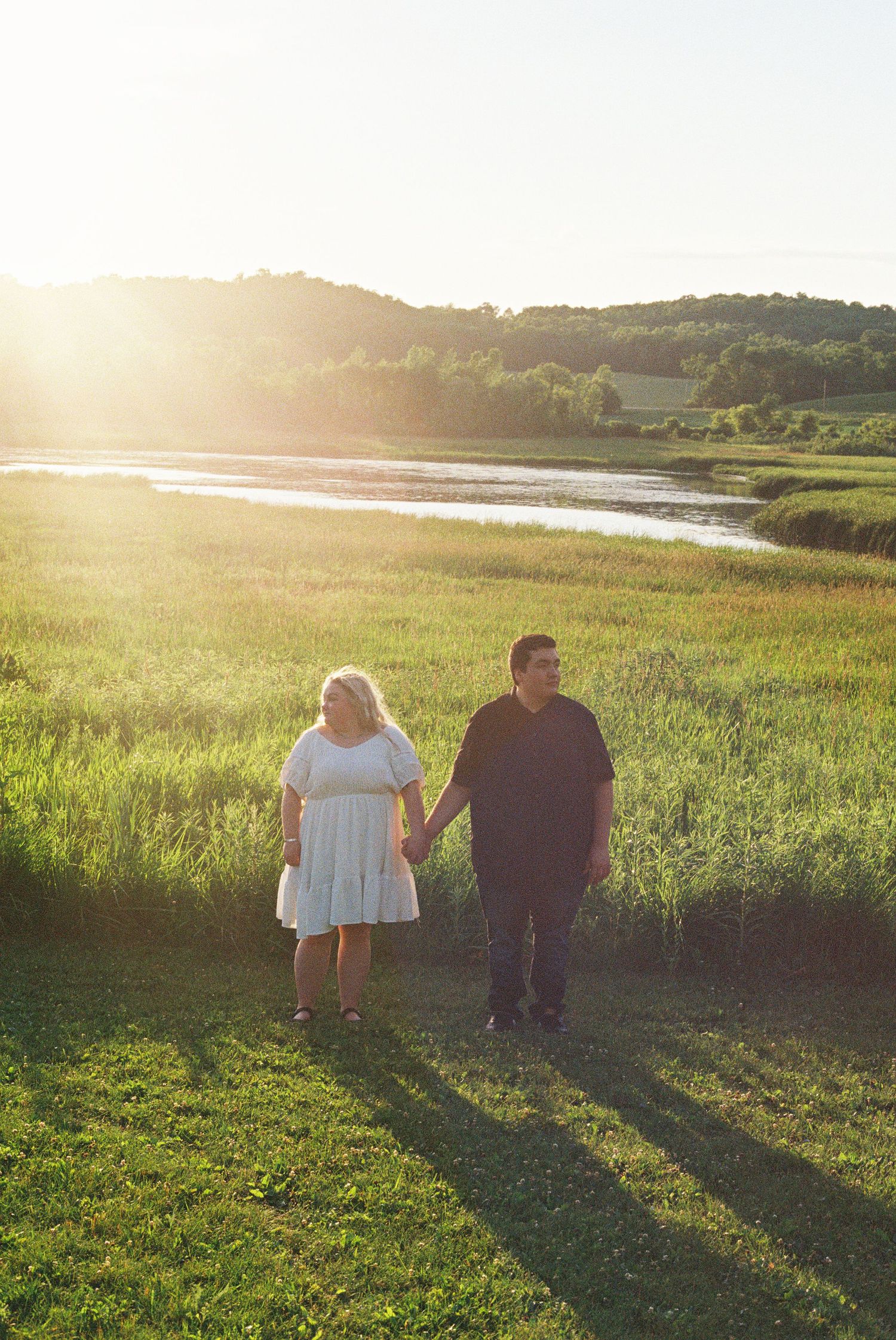 Couple holding hands walking through green meadow near river at golden sunset hour.