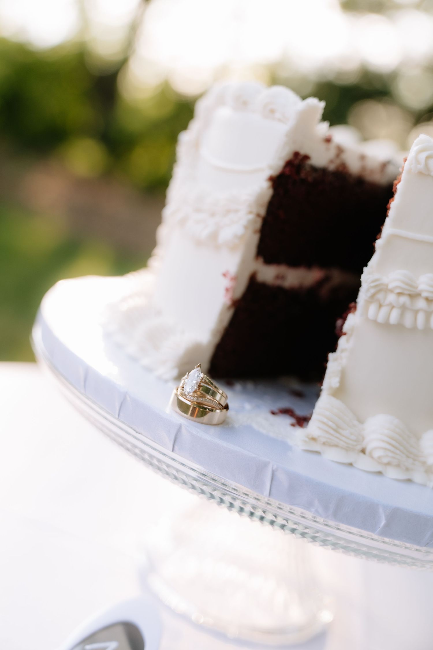 White frosted red velvet wedding cake with gold rings on cake stand, slice removed revealing chocolate layers.