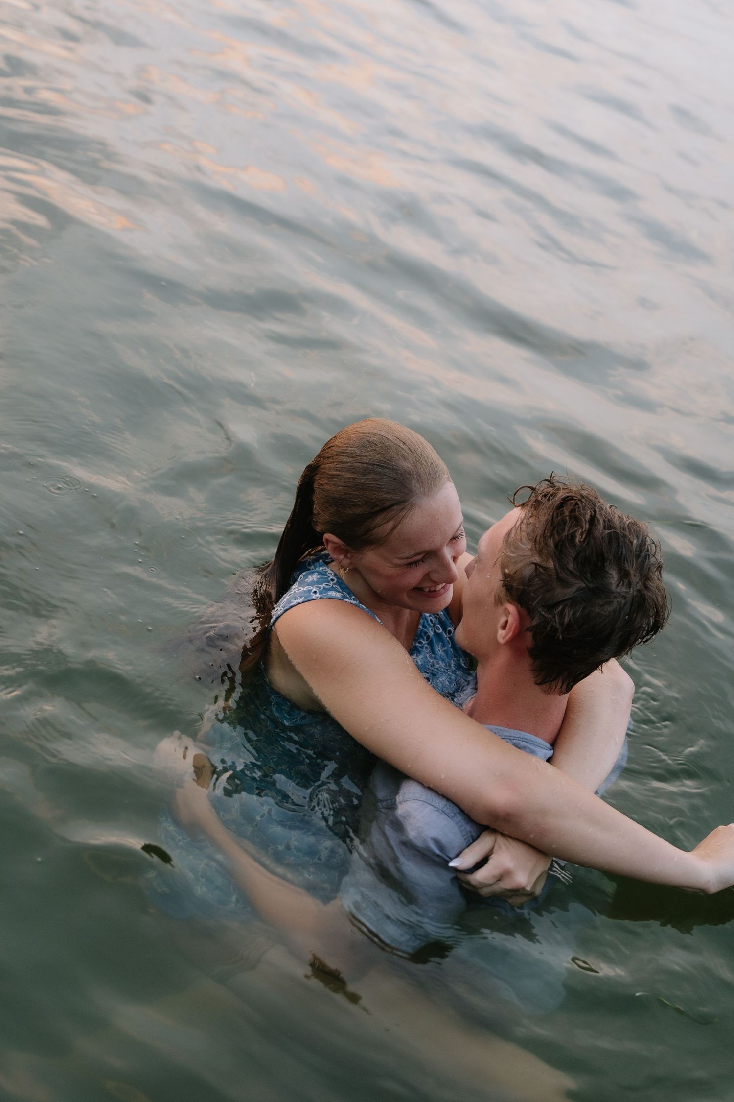 Couple embracing and smiling in a lake, wearing wet clothes, sharing a romantic moment in calm water.