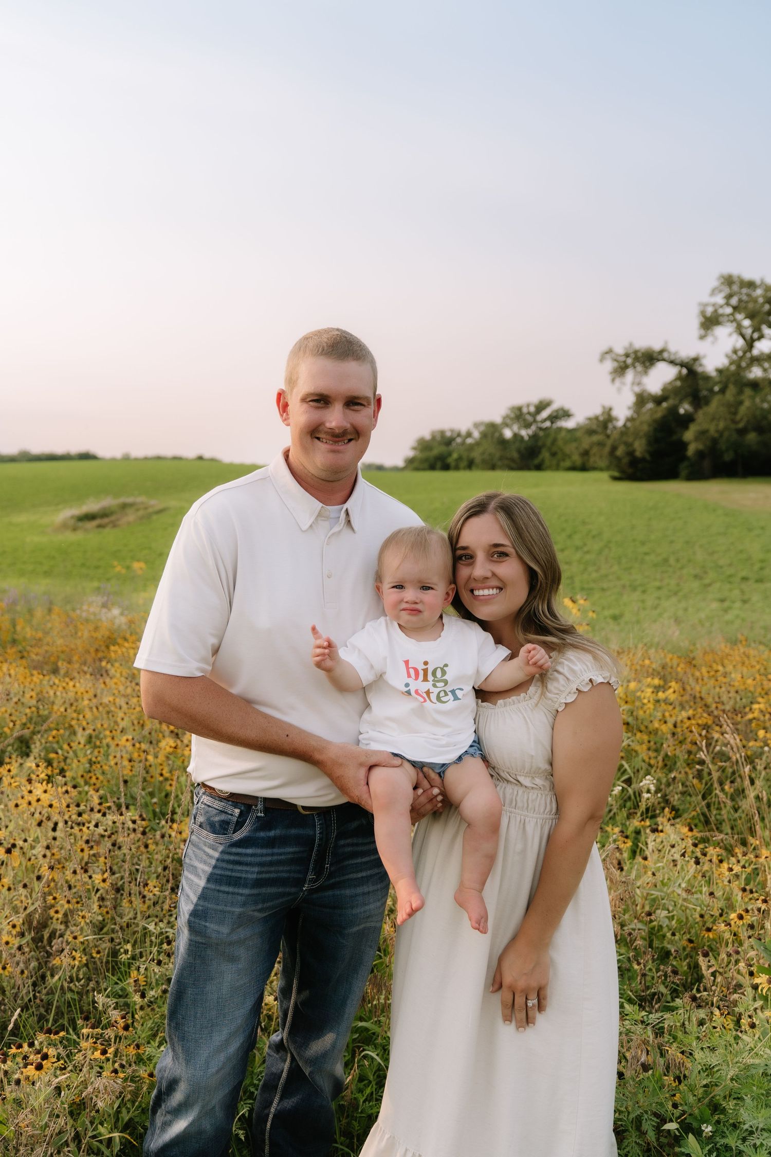A smiling family of three poses in a wildflower field, with baby wearing a big sister shirt, announcing a pregnancy.