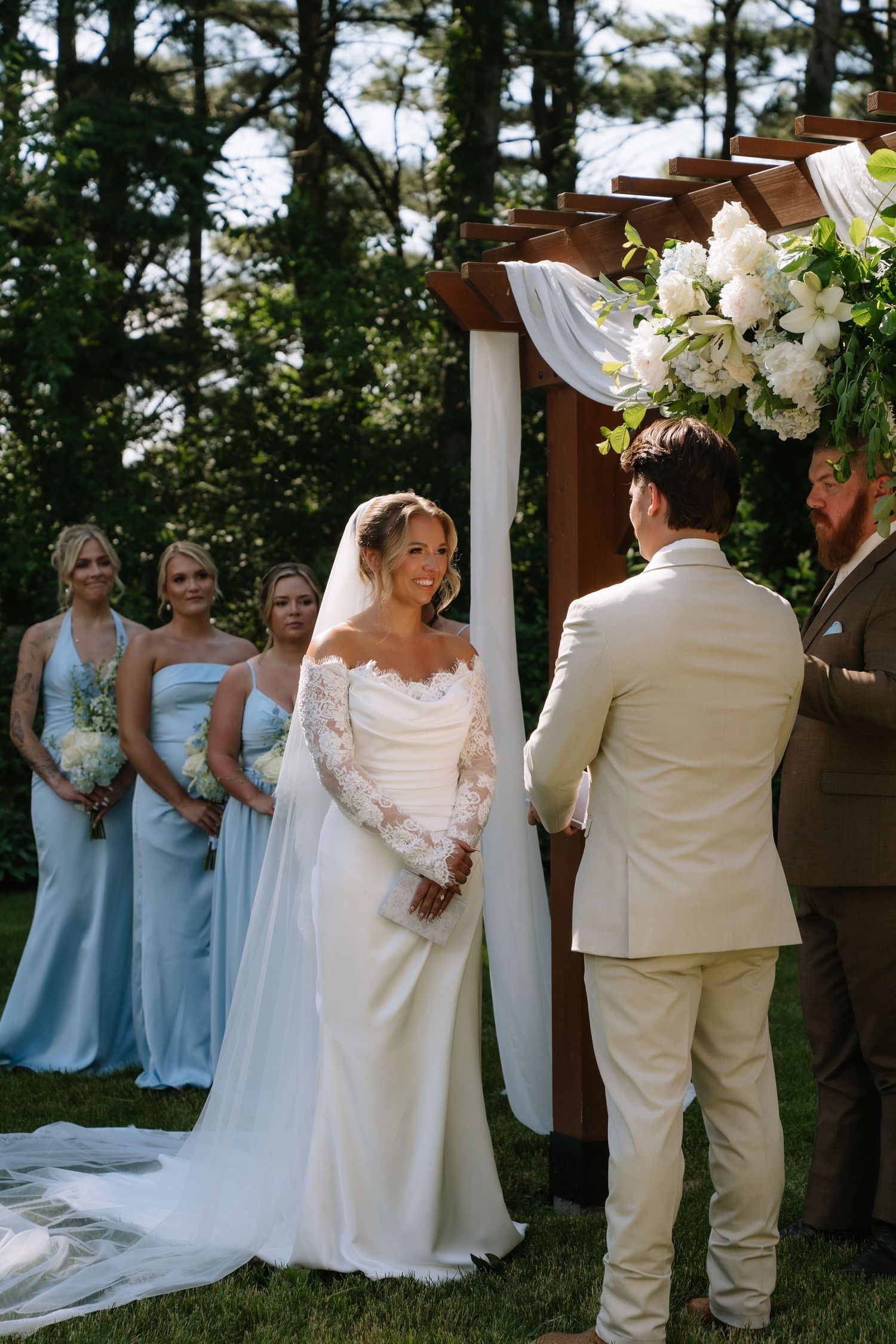 Bride in lace off-shoulder gown exchanges vows with groom in beige suit at outdoor wedding ceremony with floral arch.