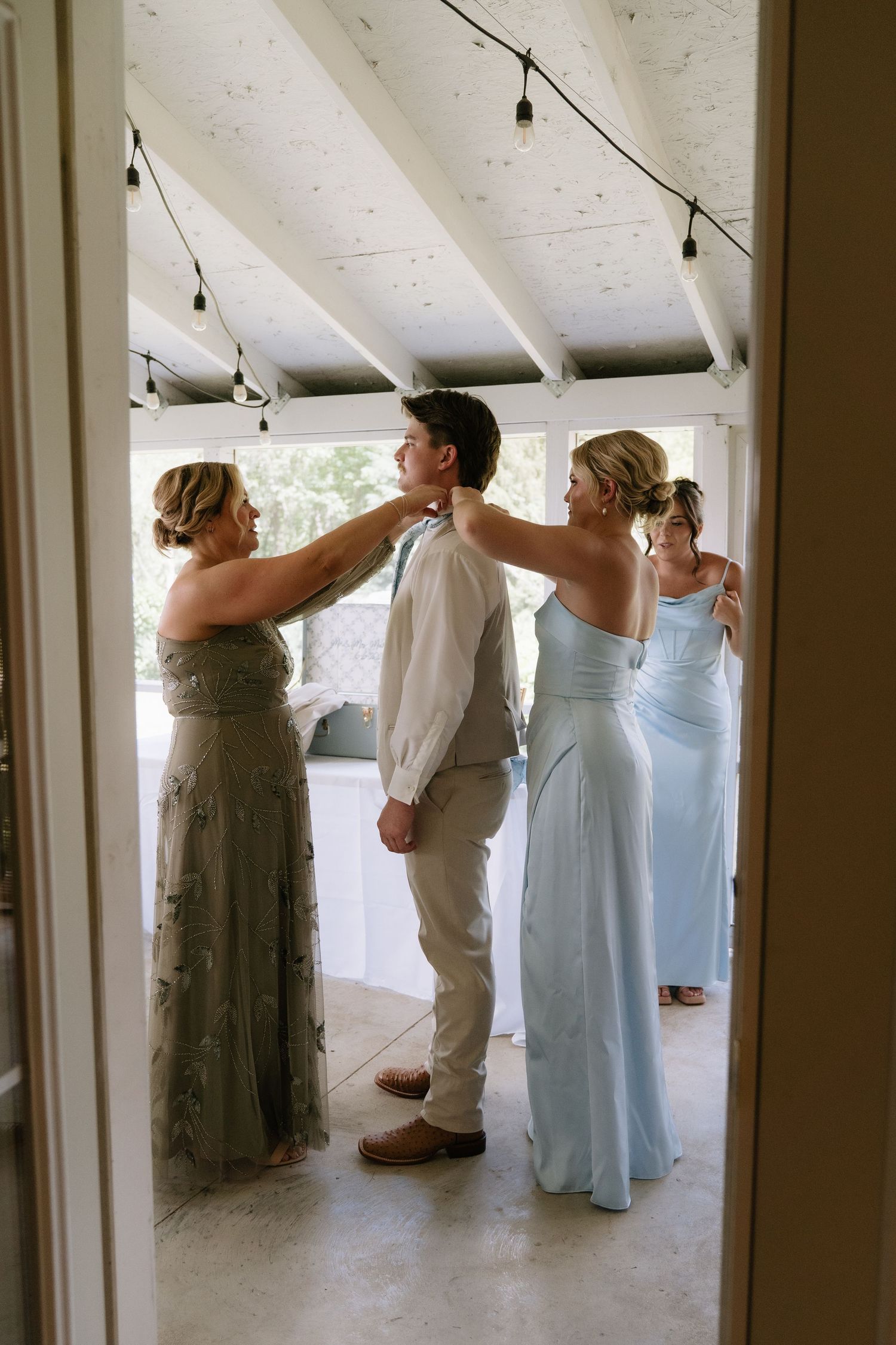 Two bridesmaids in elegant gowns help a groom adjust his tie before a wedding ceremony indoors.