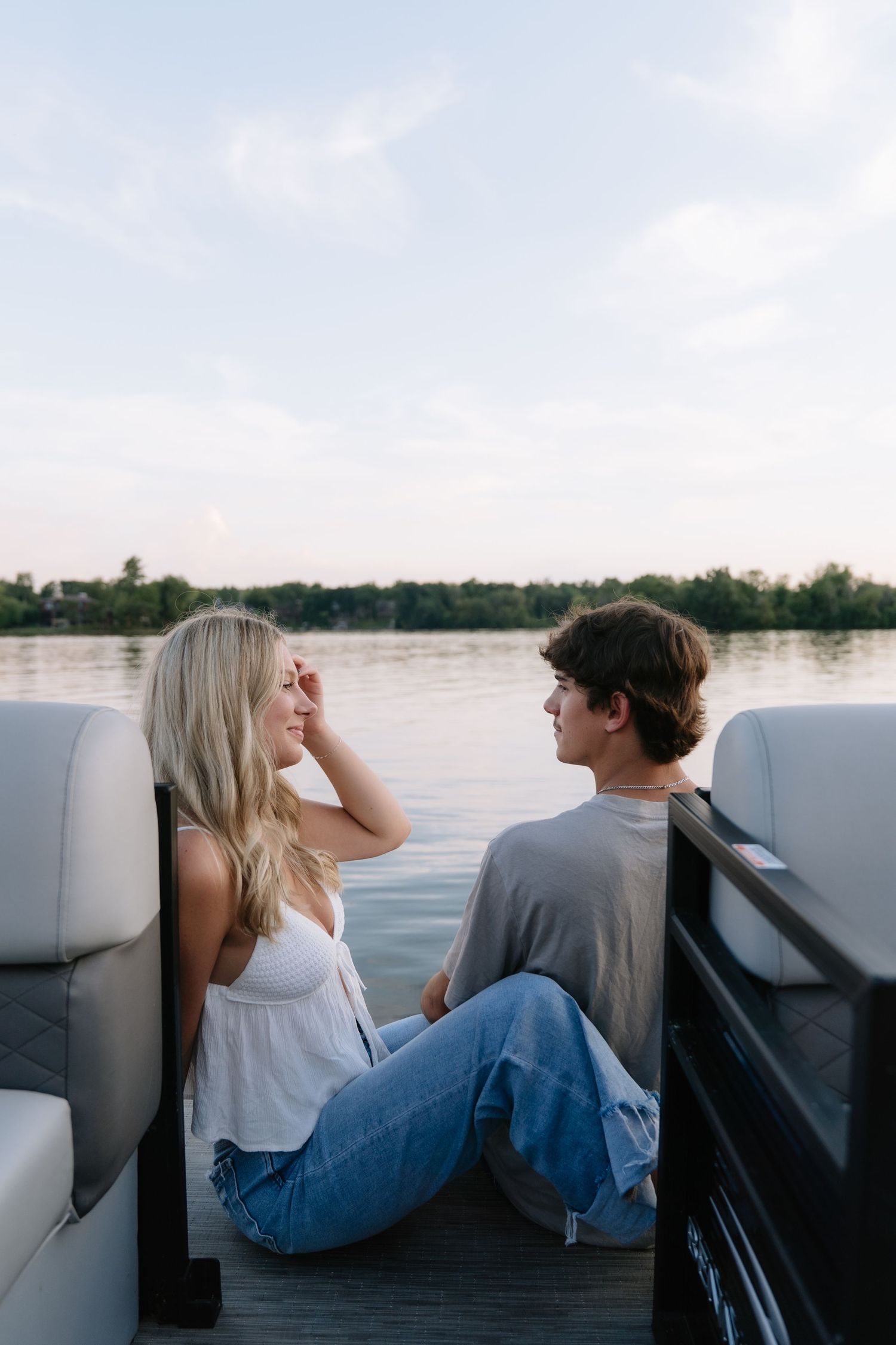 A young couple sits on a boat deck, gazing at each other with a serene lake and trees in the background.