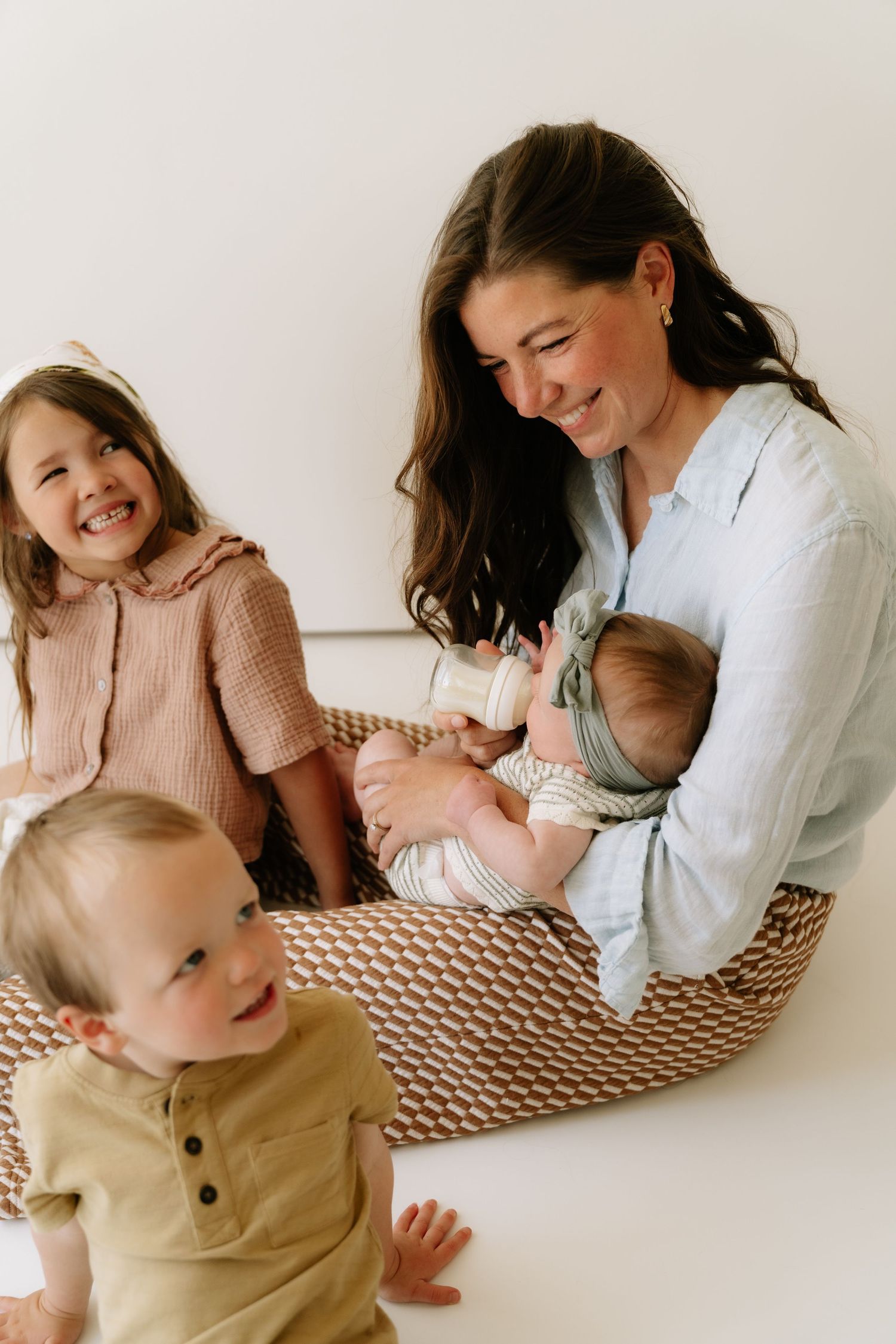A smiling mother holds a newborn while two toddlers sit nearby on a woven floor cushion in a bright room.