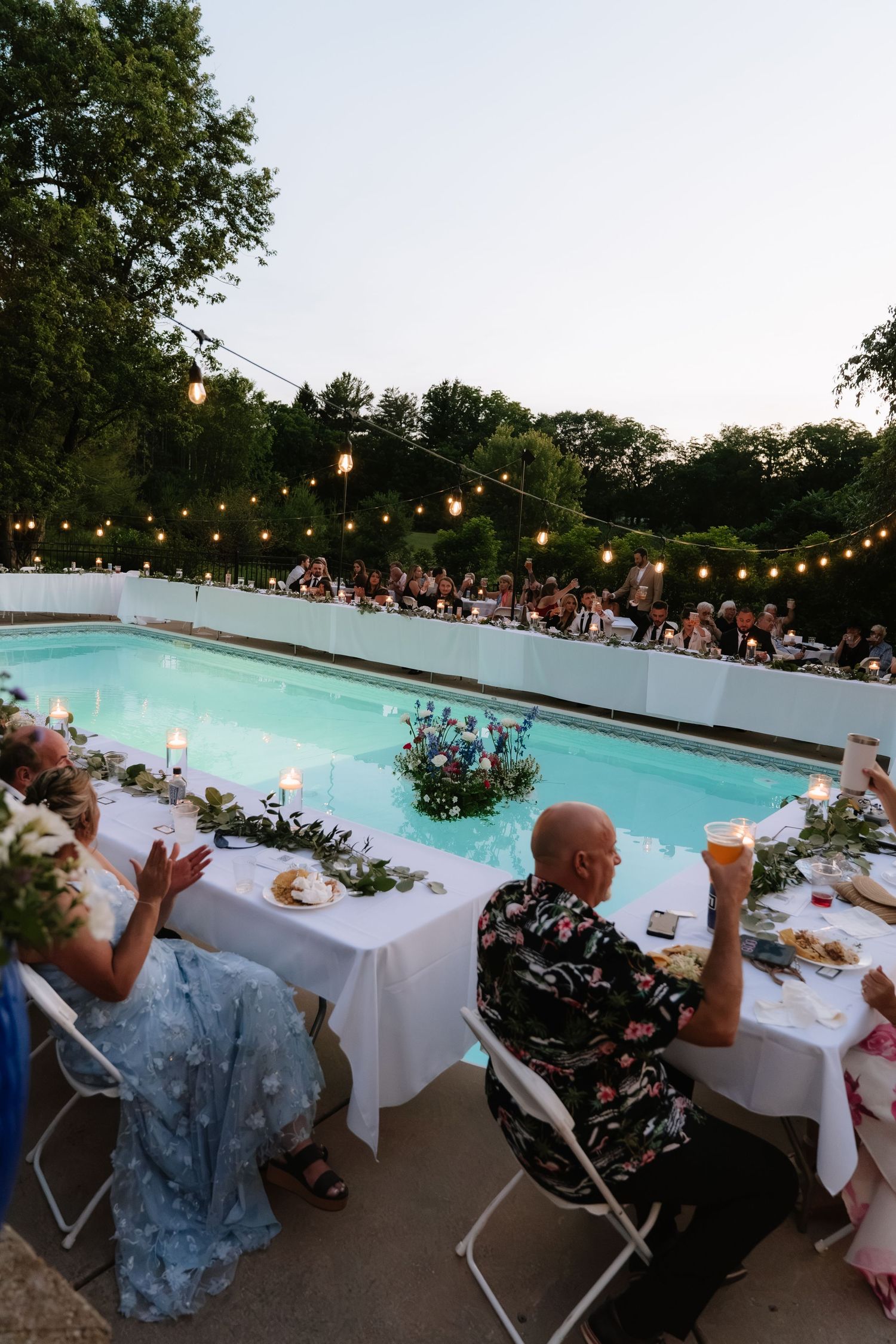 Outdoor wedding reception by a pool with string lights, guests dining at white tables adorned with floral centerpieces at dusk.
