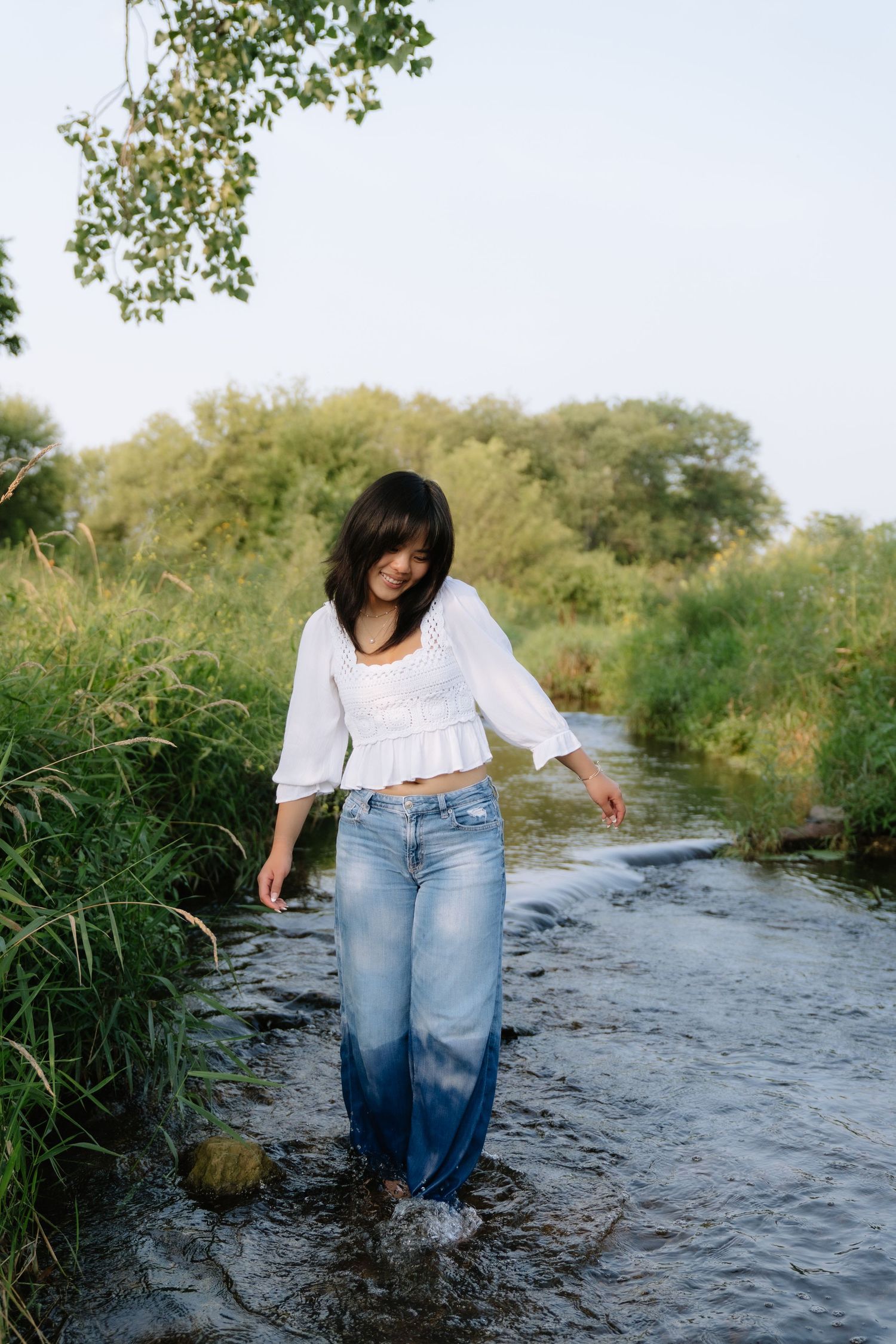 Young woman in white top and wet jeans wading through a shallow stream surrounded by lush green vegetation.