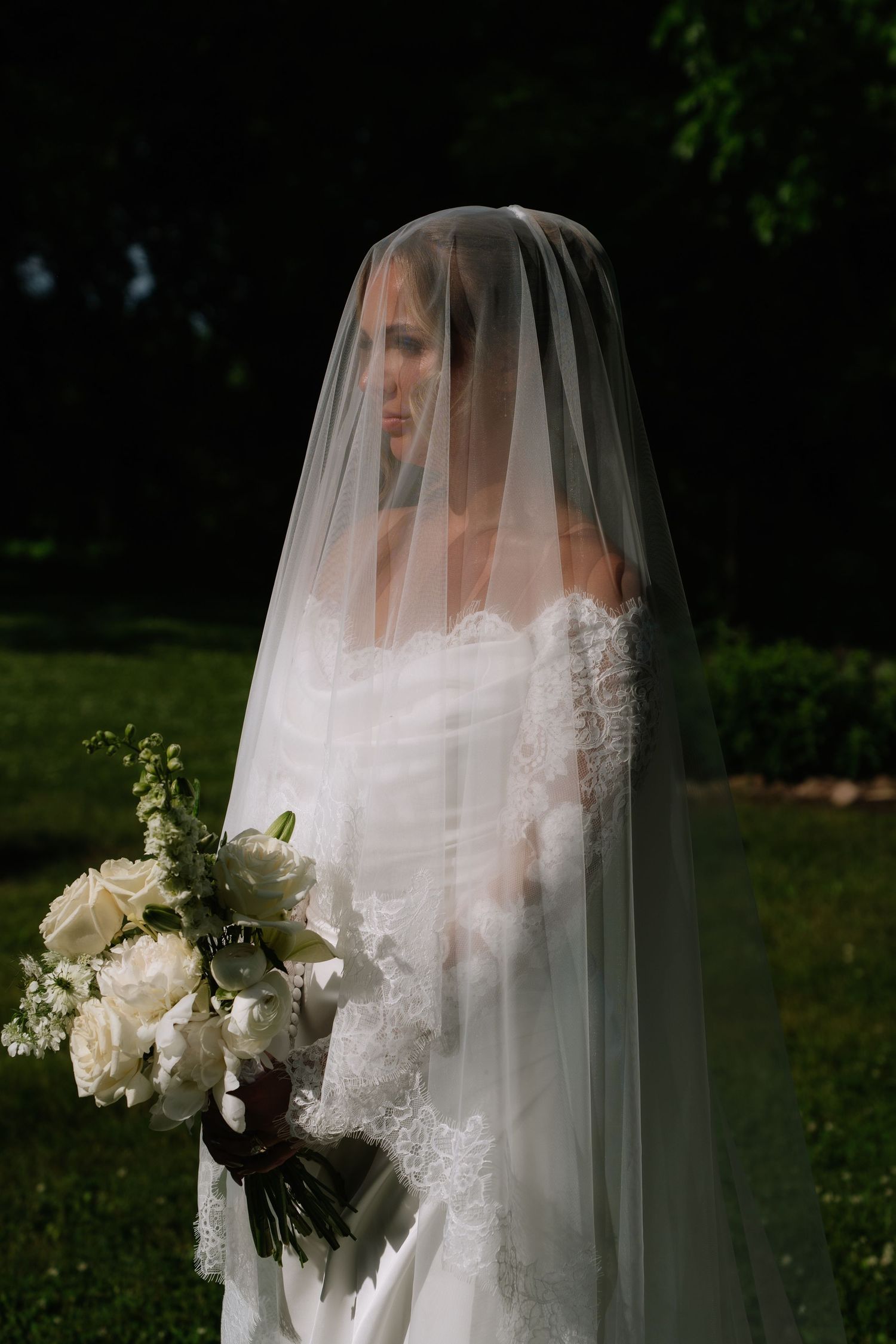 Bride wearing a lace wedding dress and sheer veil over her face, holding a white rose bouquet outdoors.