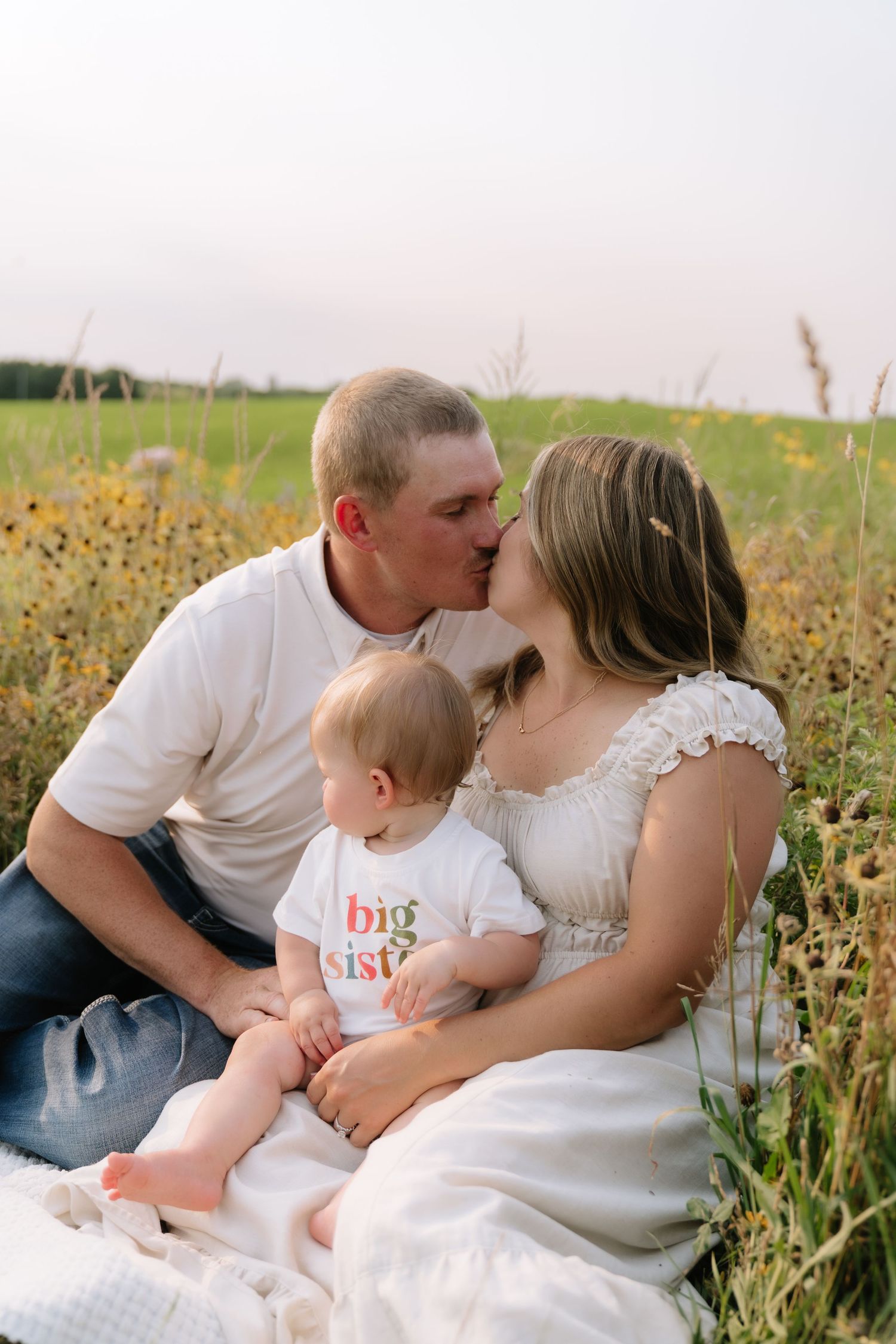 A couple kisses in a flower field while their baby in a big sister shirt sits on mom's lap.