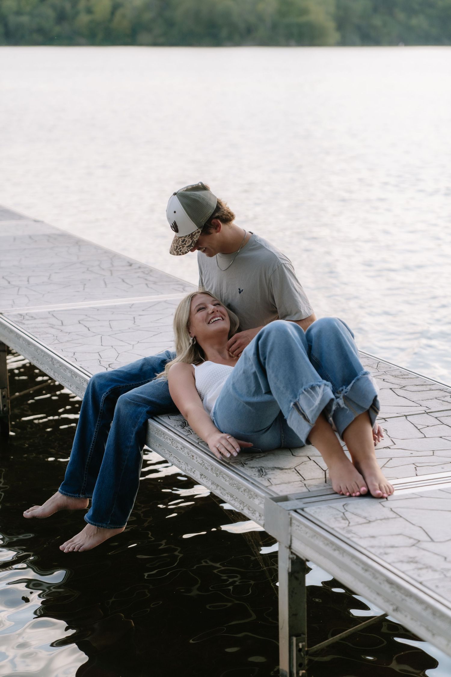 Couple sitting on a lakeside dock, woman leaning back laughing, man in cap smiling down at her, barefoot in jeans.