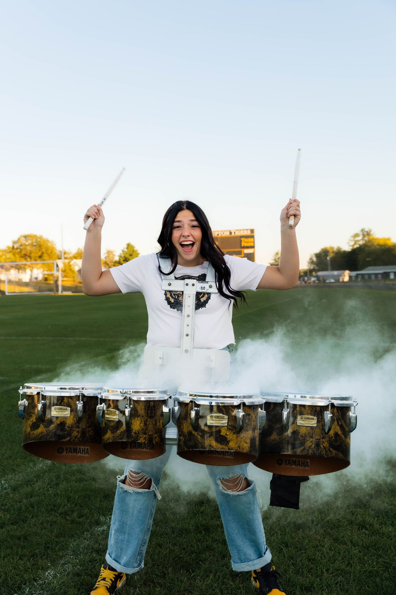 Excited female drummer raises sticks above Yamaha tenor drums with dramatic smoke effect on field.