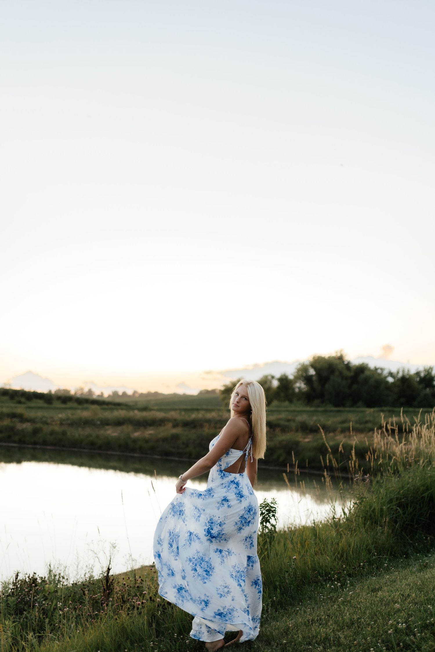 Blonde girl in white and blue floral maxi dress standing by a pond at sunset in a rural field.