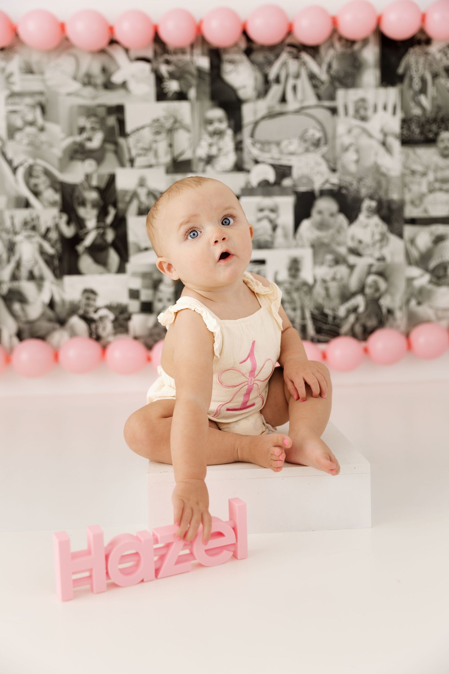 Baby girl named Hazel sits on white block beside pink name sign with photo collage backdrop.