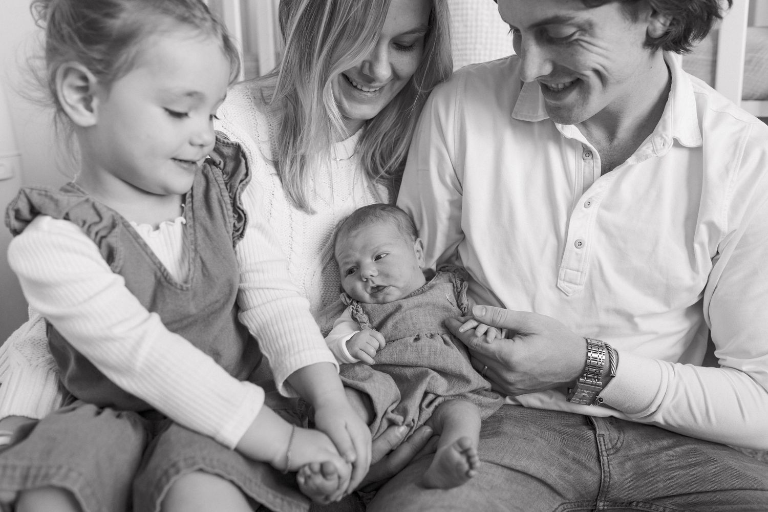 Black and white photo of a happy family of four admiring their newborn baby together.