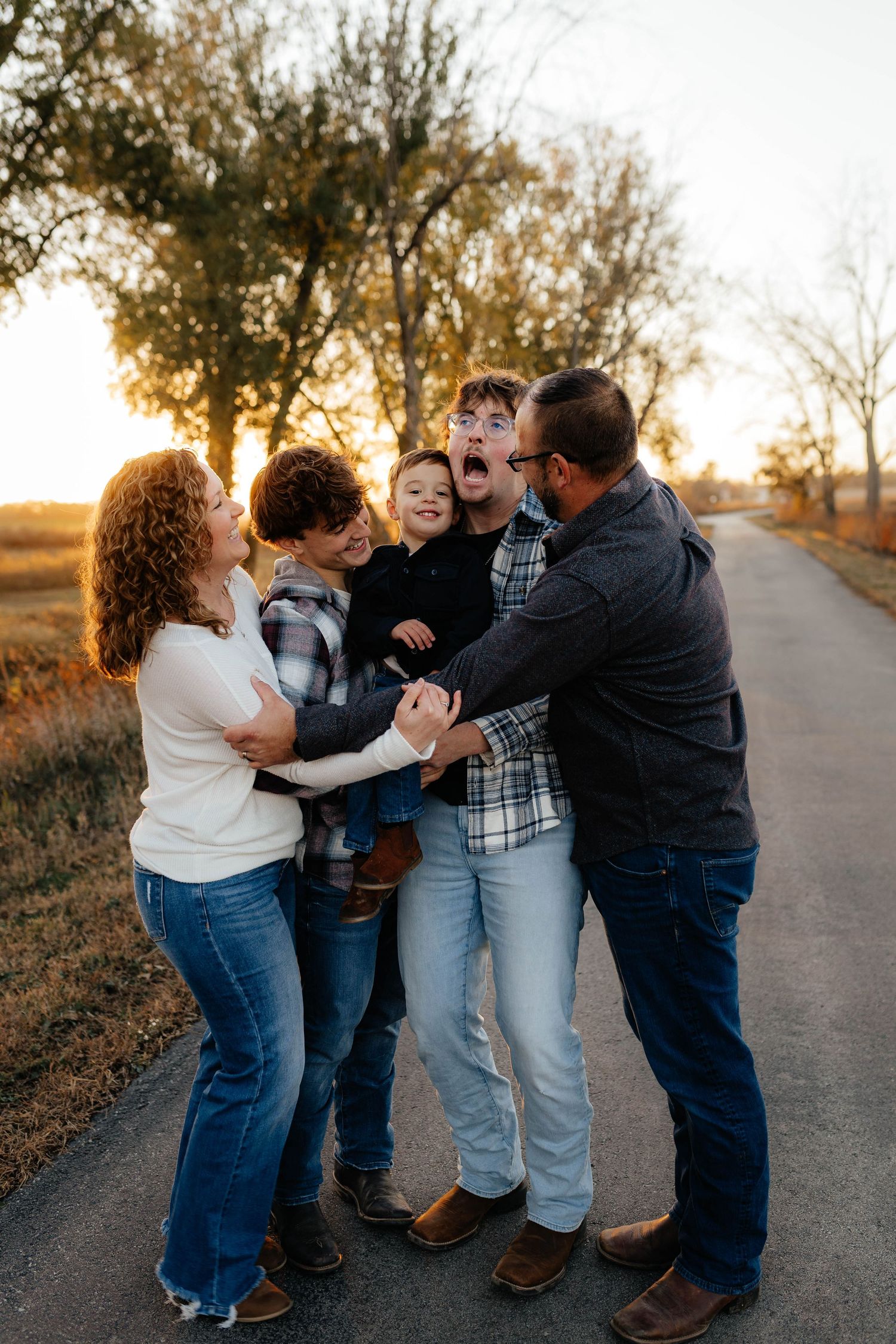 Joyful family of five hugging and laughing together on rural road at sunset.