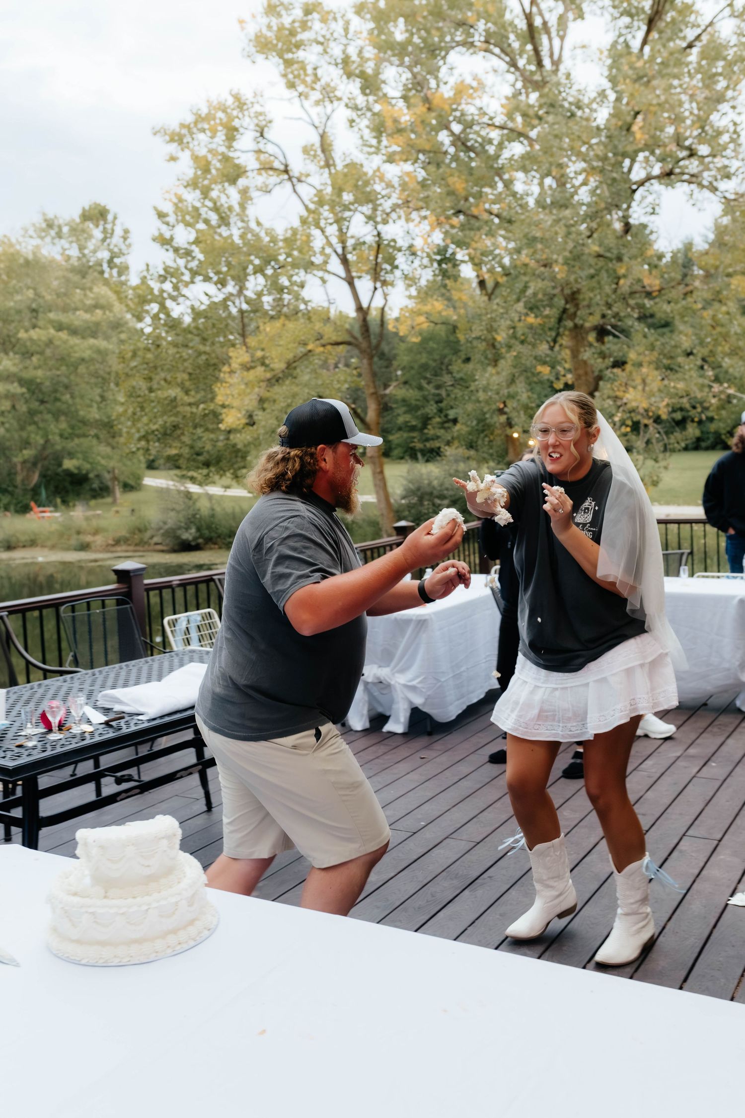 Bride and groom laughing while playfully smearing wedding cake on an outdoor deck.