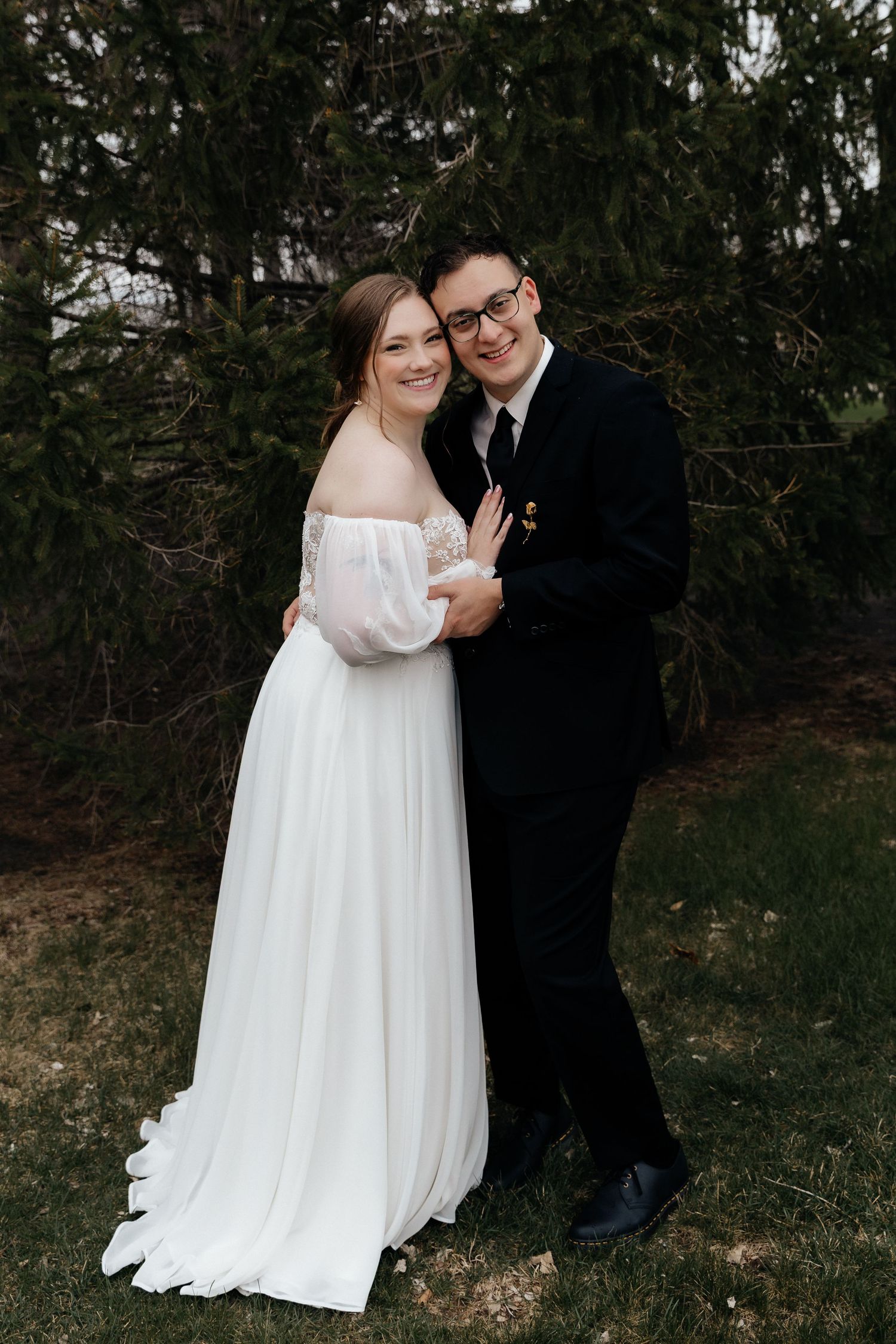 Smiling bride and groom embracing outdoors in front of evergreen trees, bride wearing off-shoulder lace wedding gown.