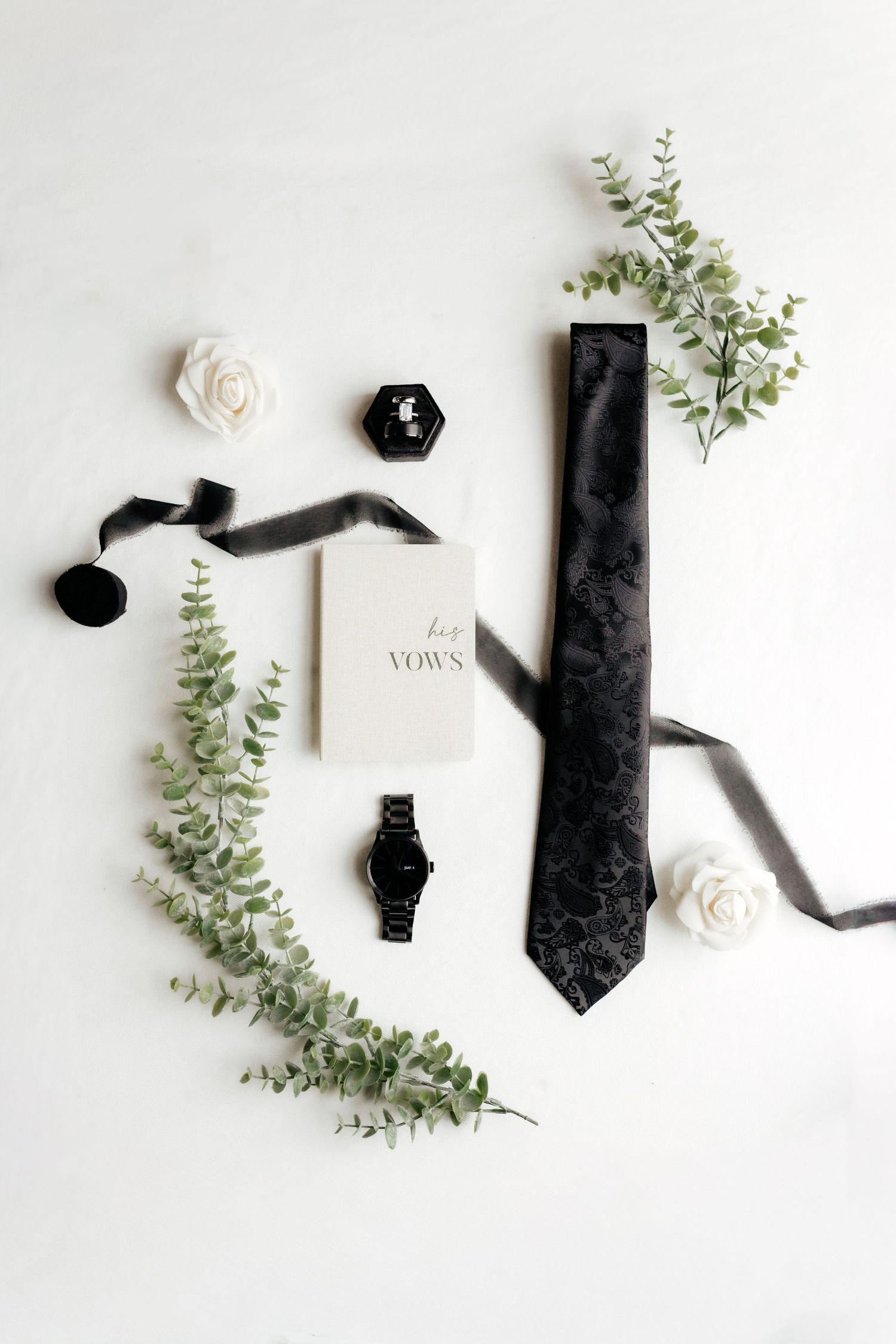 Groom flat lay with black tie, vow book, watch, ring box, roses, and eucalyptus on white background.