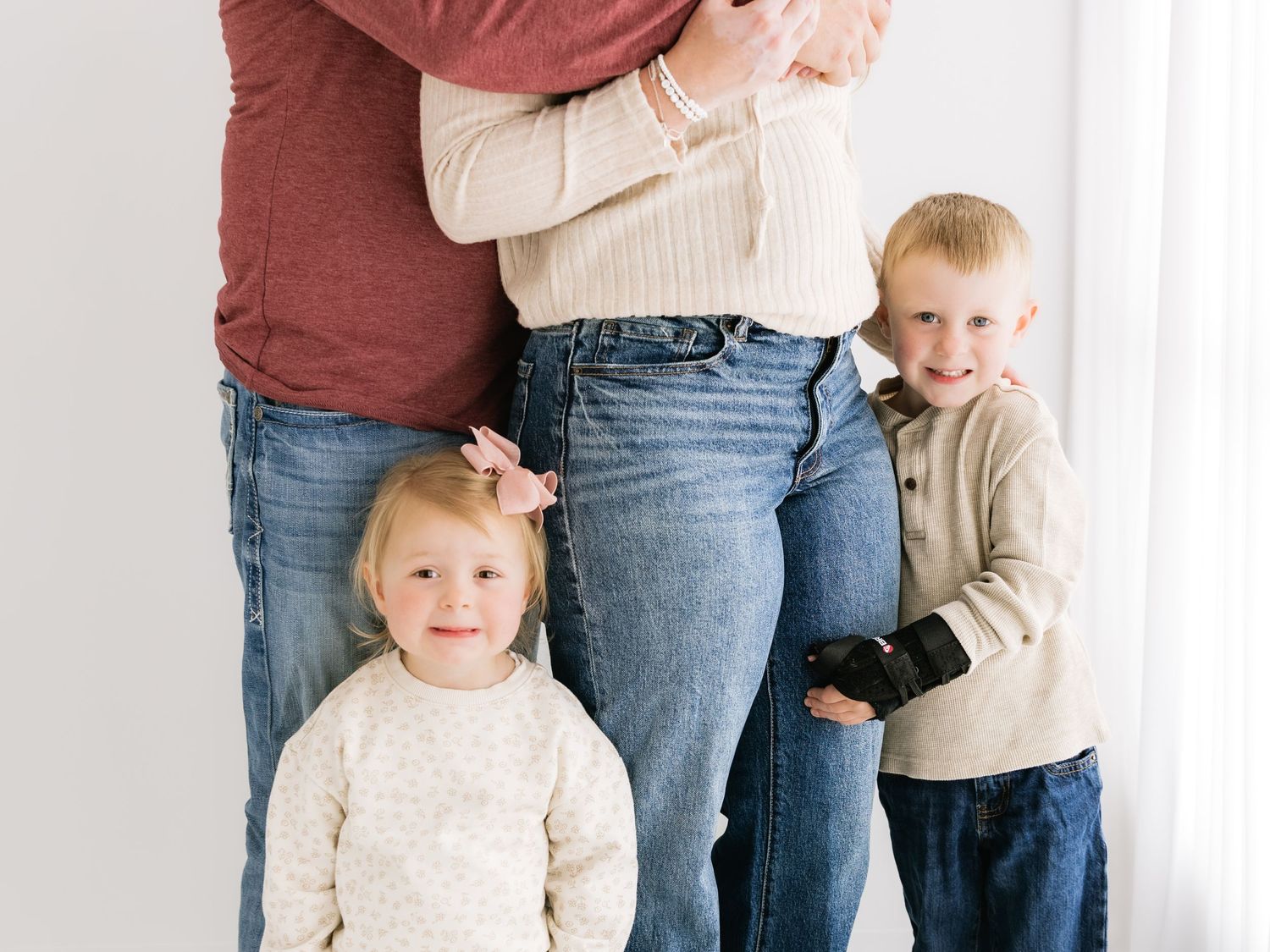 Family of four posing together, parents embracing while two young children smile at camera in bright white studio.