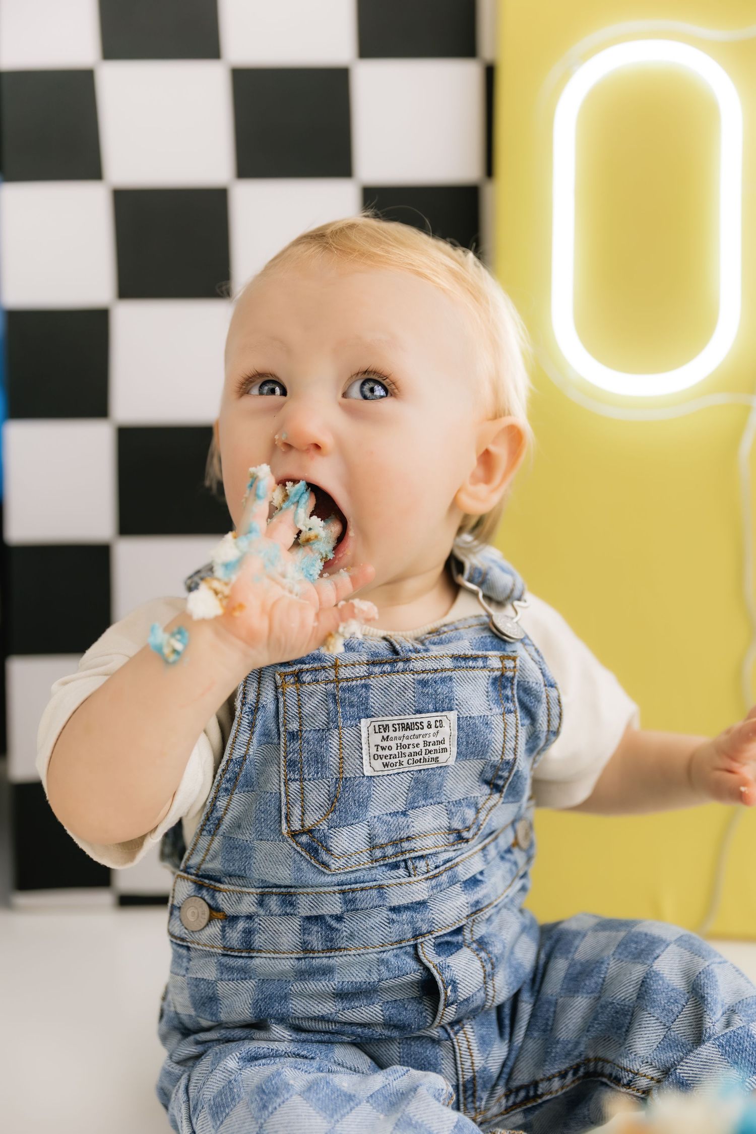 Adorable blue-eyed baby in denim overalls eating birthday cake frosting at first birthday cake smash session.