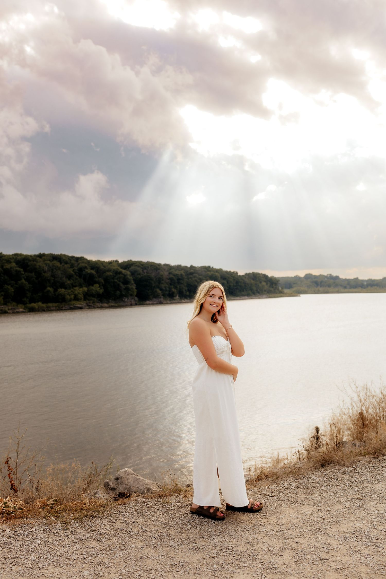 Blonde woman in white dress standing by a lake with dramatic sunbeams breaking through clouds.