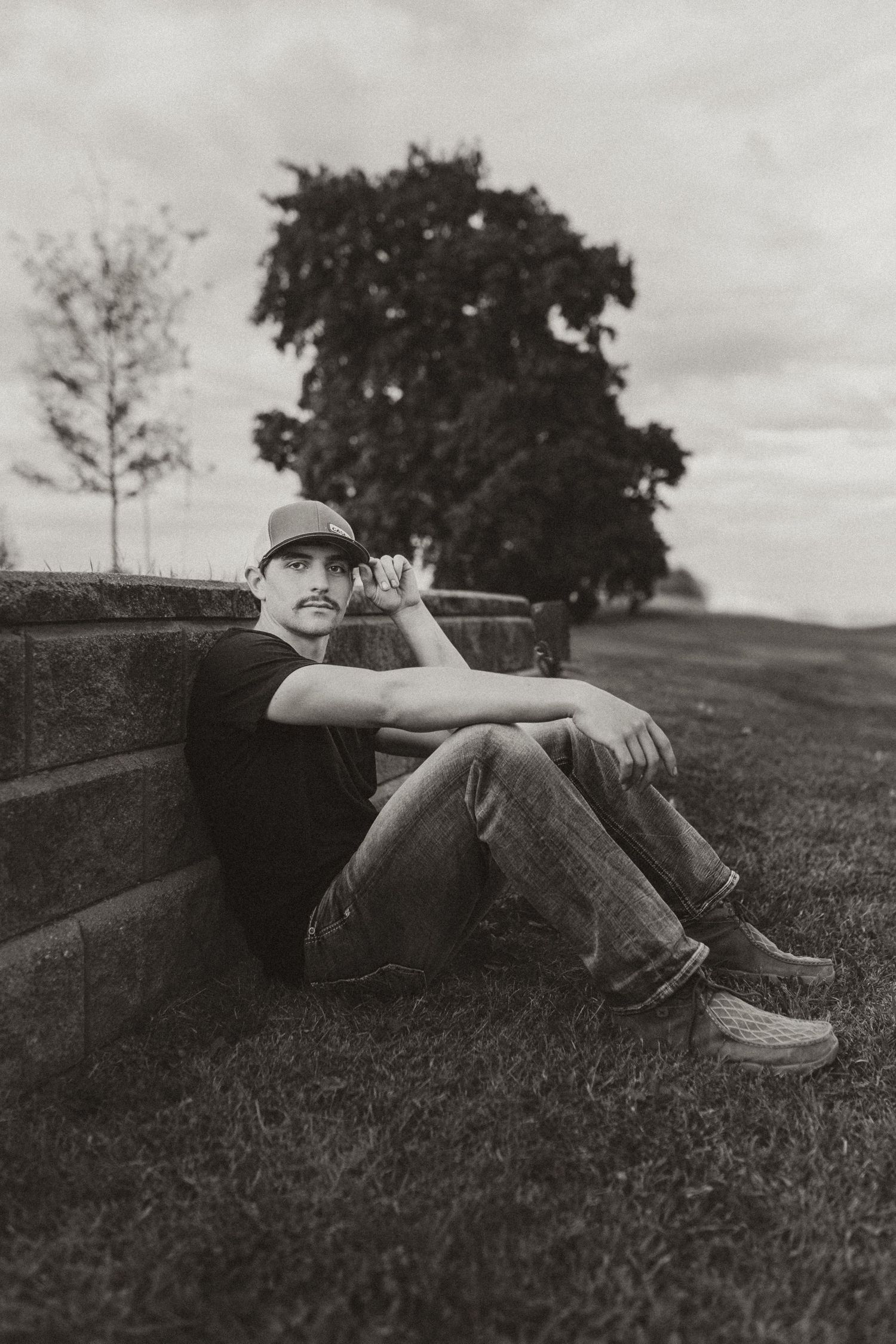 Black and white photo of a young man in a hat sitting against a stone wall in a park setting.