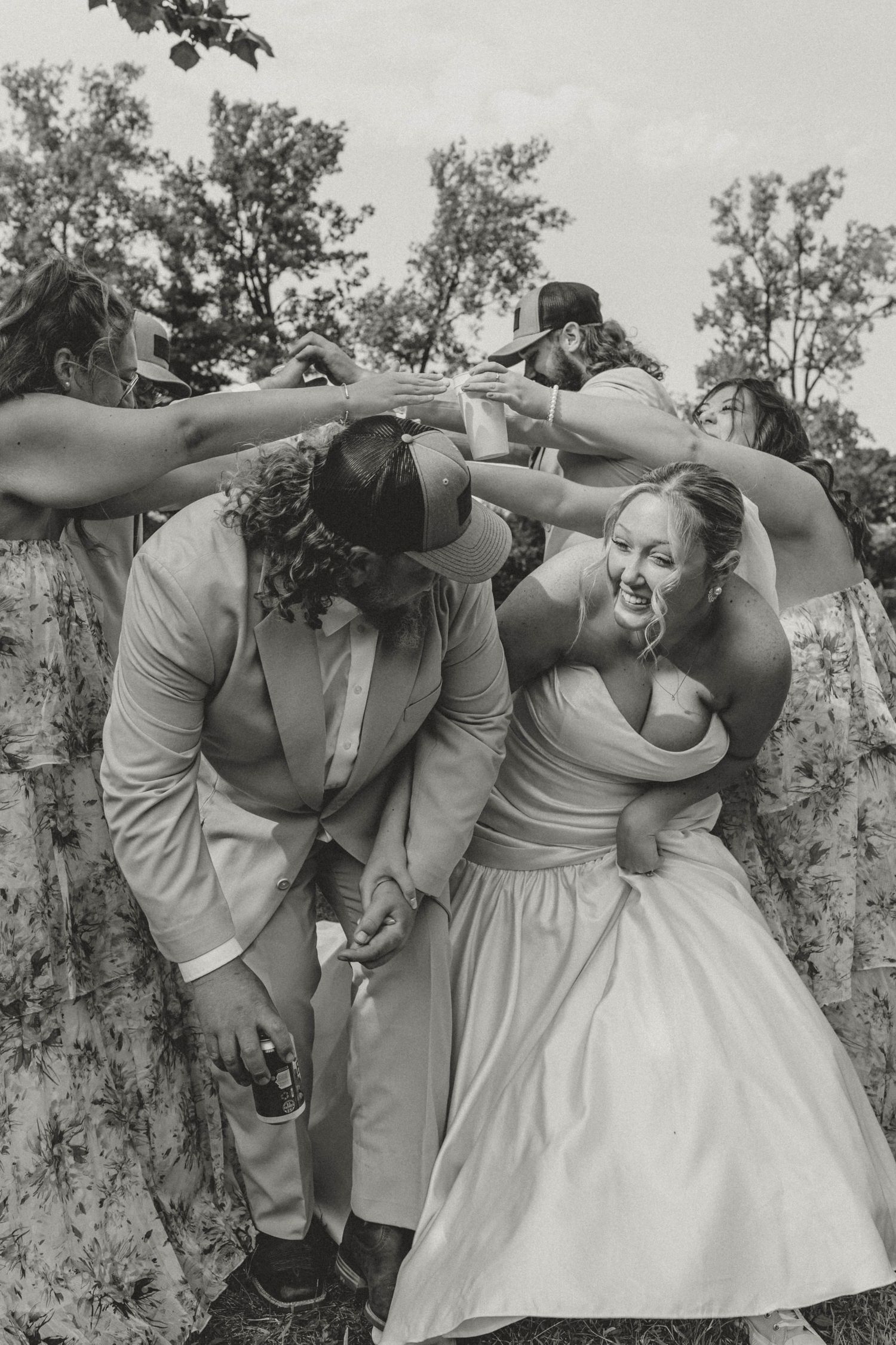Black and white photo of bride and groom celebrating joyfully with wedding party under raised arms outdoors.