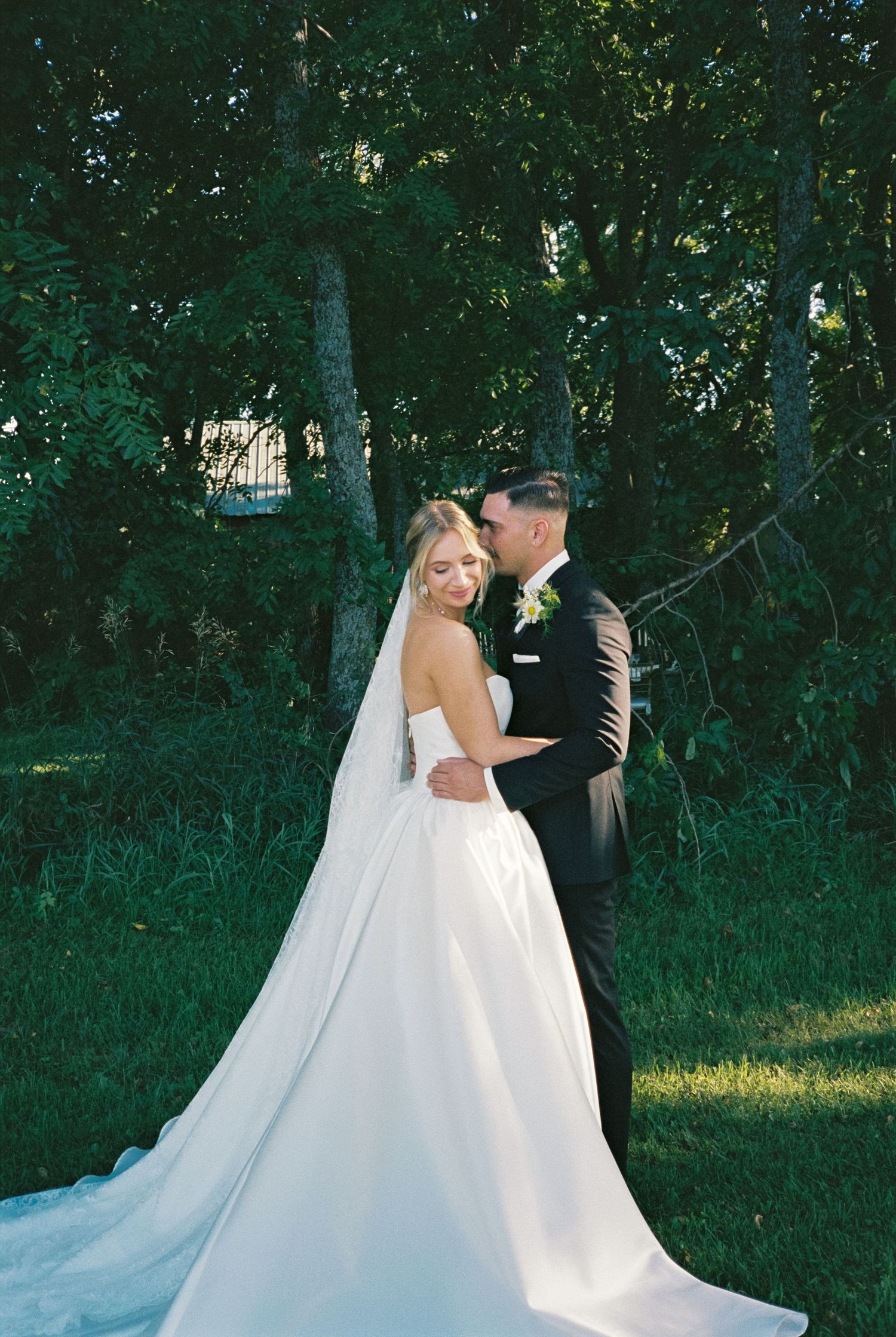 Bride and groom share a romantic embrace outdoors, surrounded by lush green trees on their wedding day.