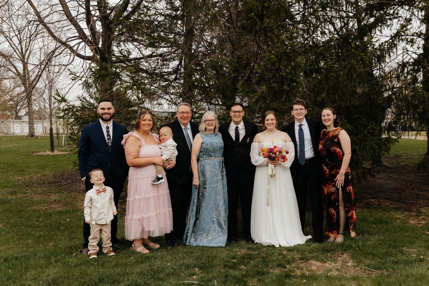 Large wedding party group photo on green lawn with bride holding vibrant bouquet.