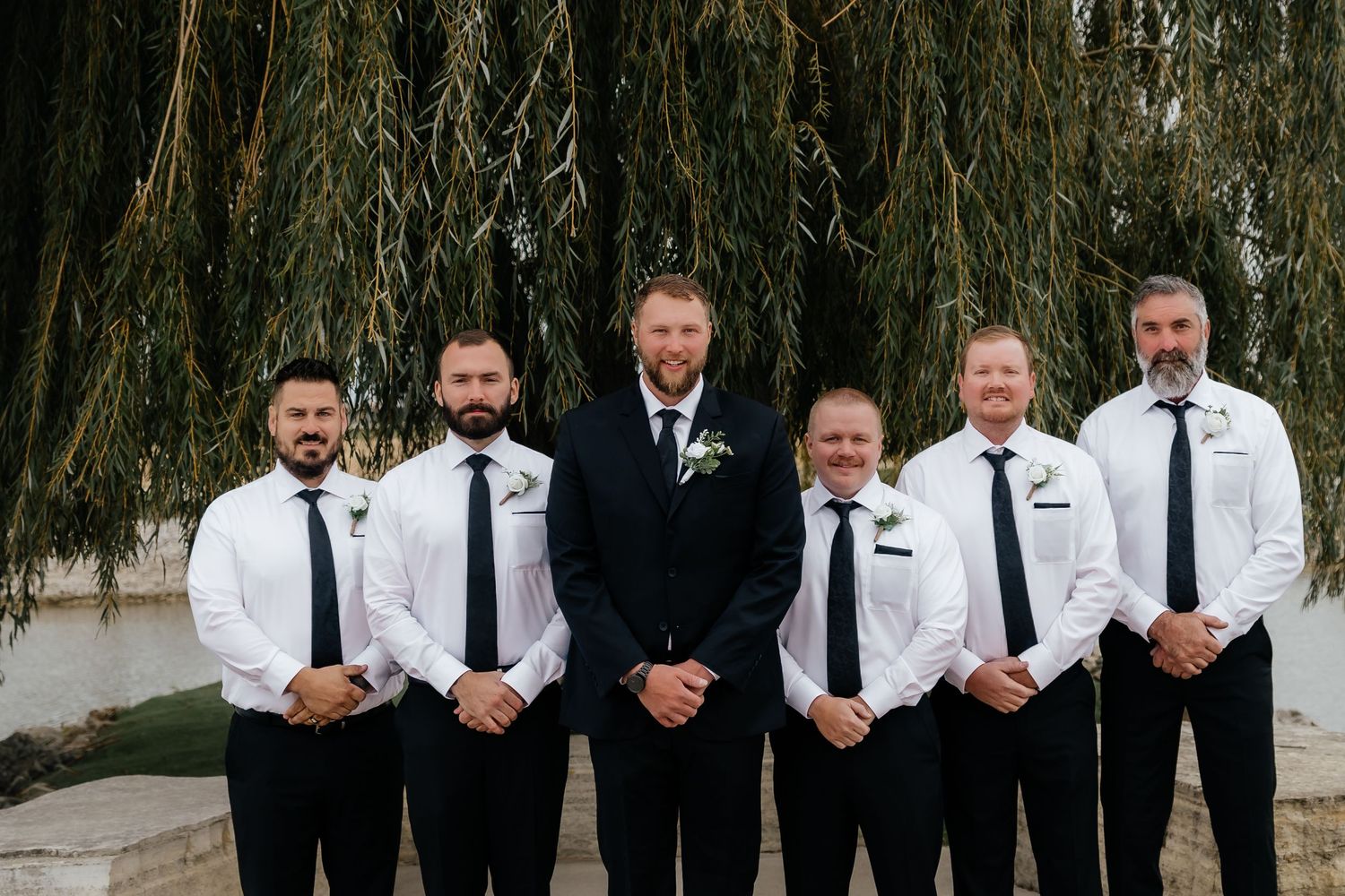 Groom in black suit standing with five groomsmen in white shirts under willow tree.