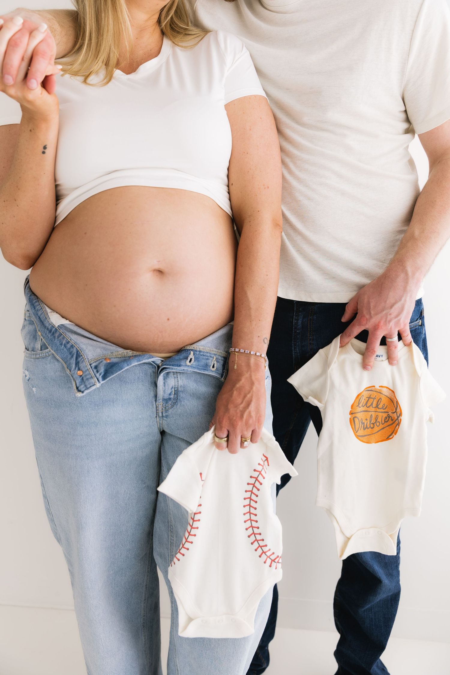 Expecting couple holds baseball and basketball themed baby onesies beside pregnant belly in studio.