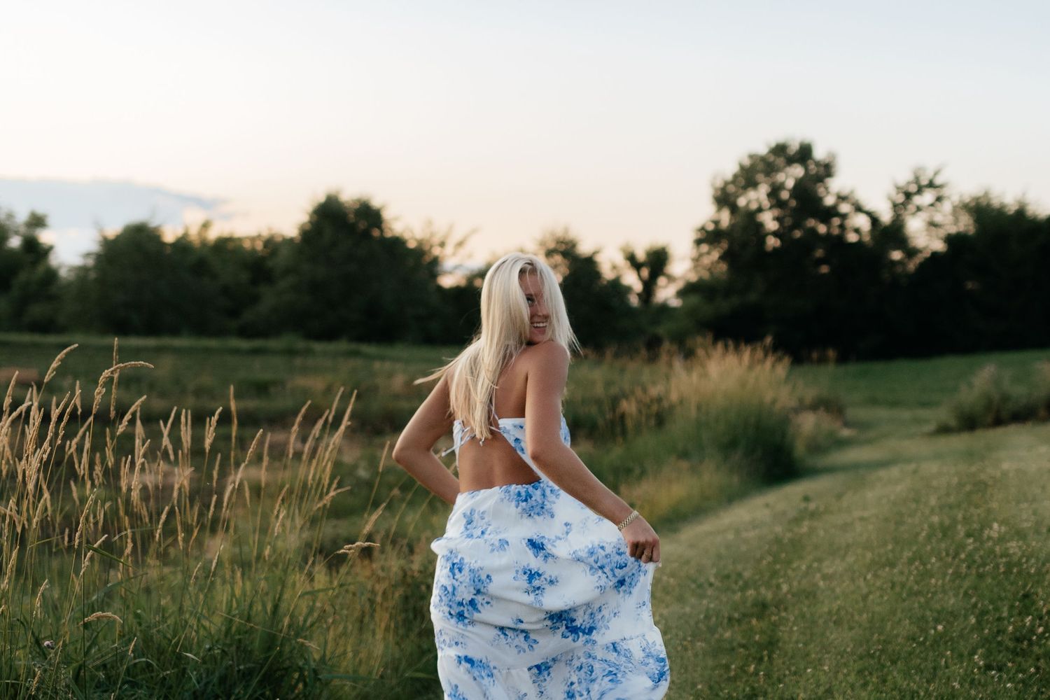 Blonde girl in white crop top and blue floral skirt walking through a lush green field at dusk.