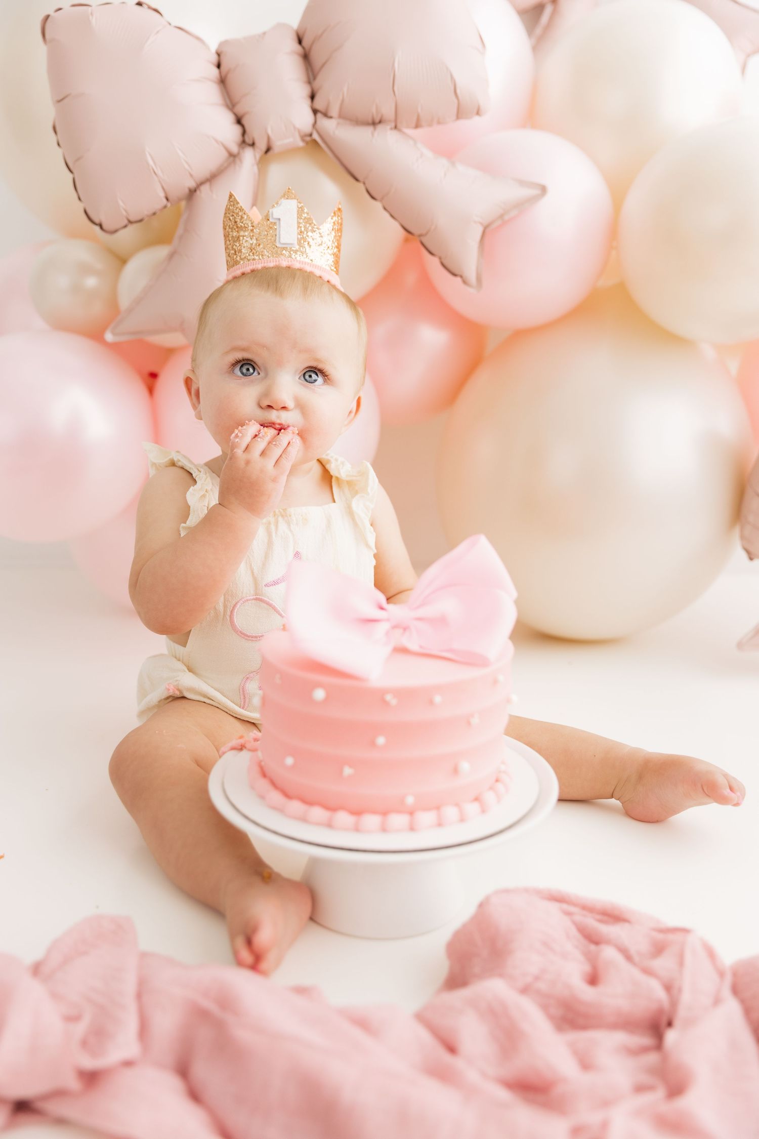 Baby girl wearing gold crown sits beside decorated pink polka dot cake with bow topper.