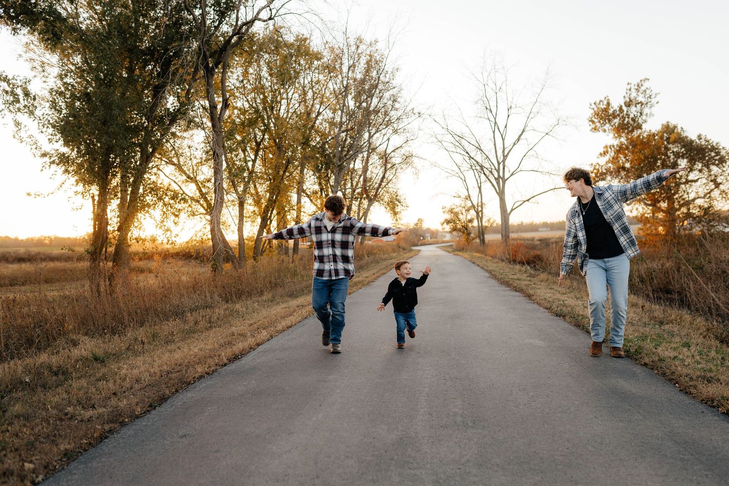 Family of three running joyfully with arms out on tree-lined road at sunset.