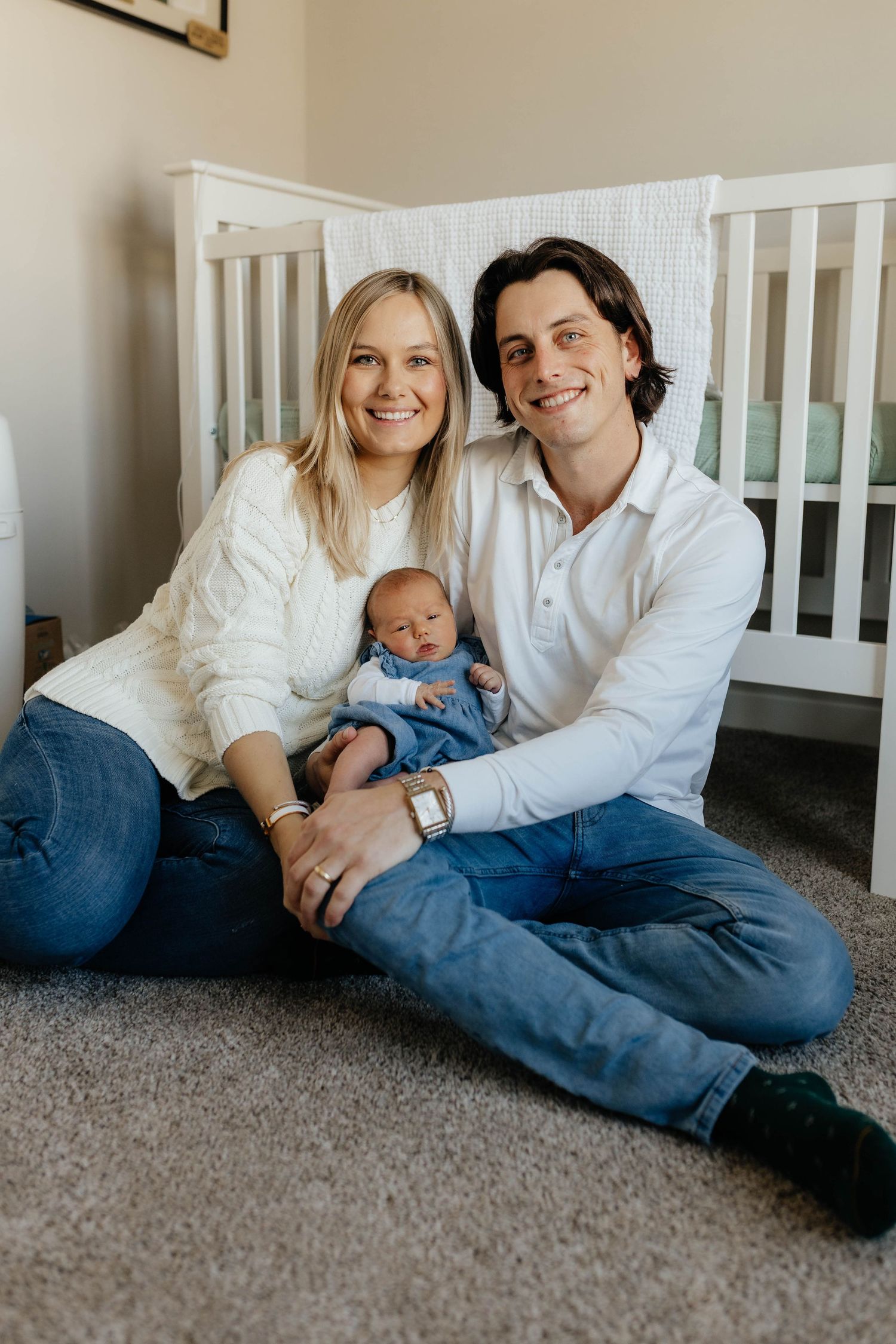Smiling parents sit on a nursery floor in front of a white crib, holding their newborn baby.