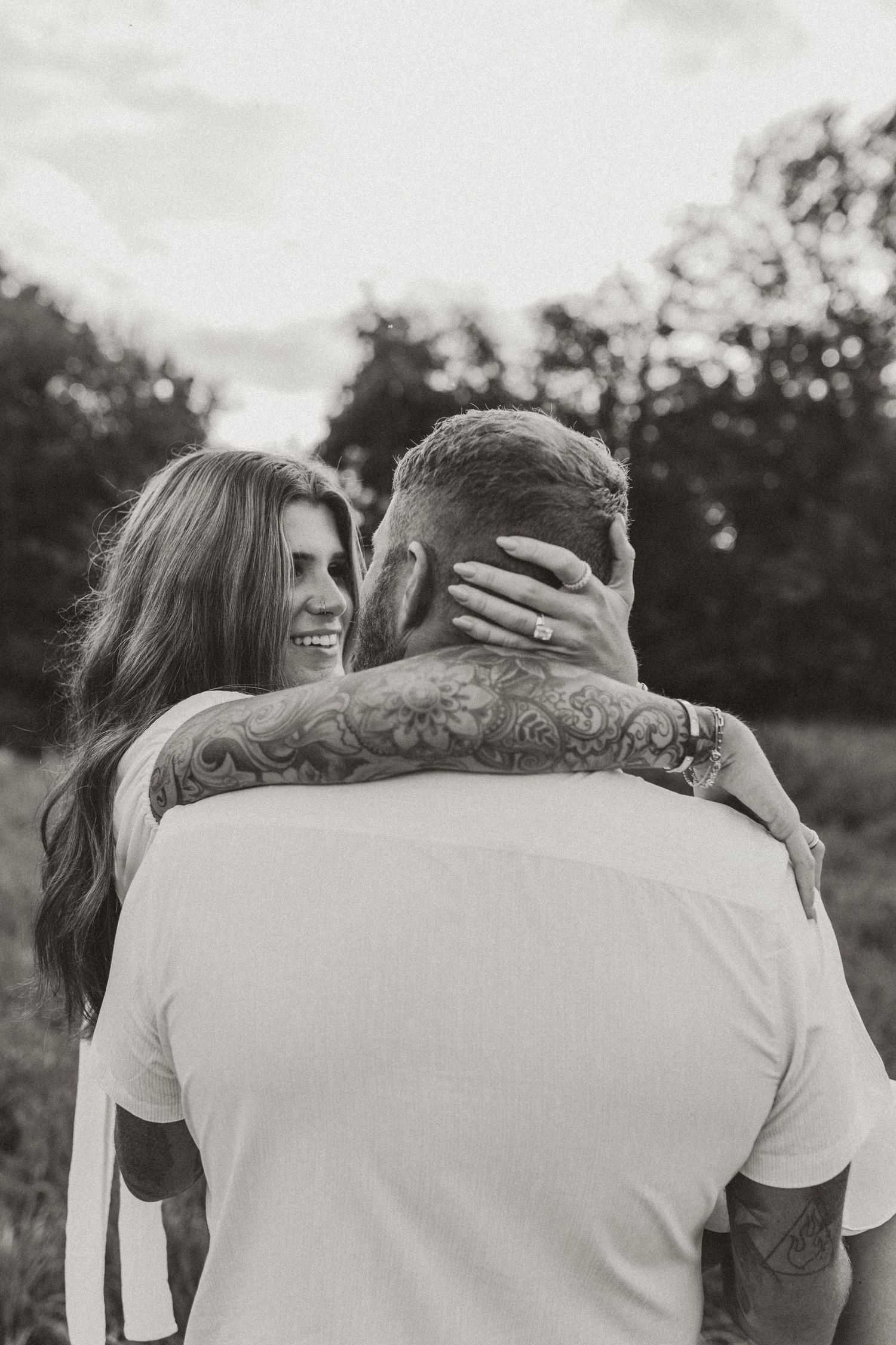 Black and white photo of a smiling woman embracing a tattooed man outdoors in a field.