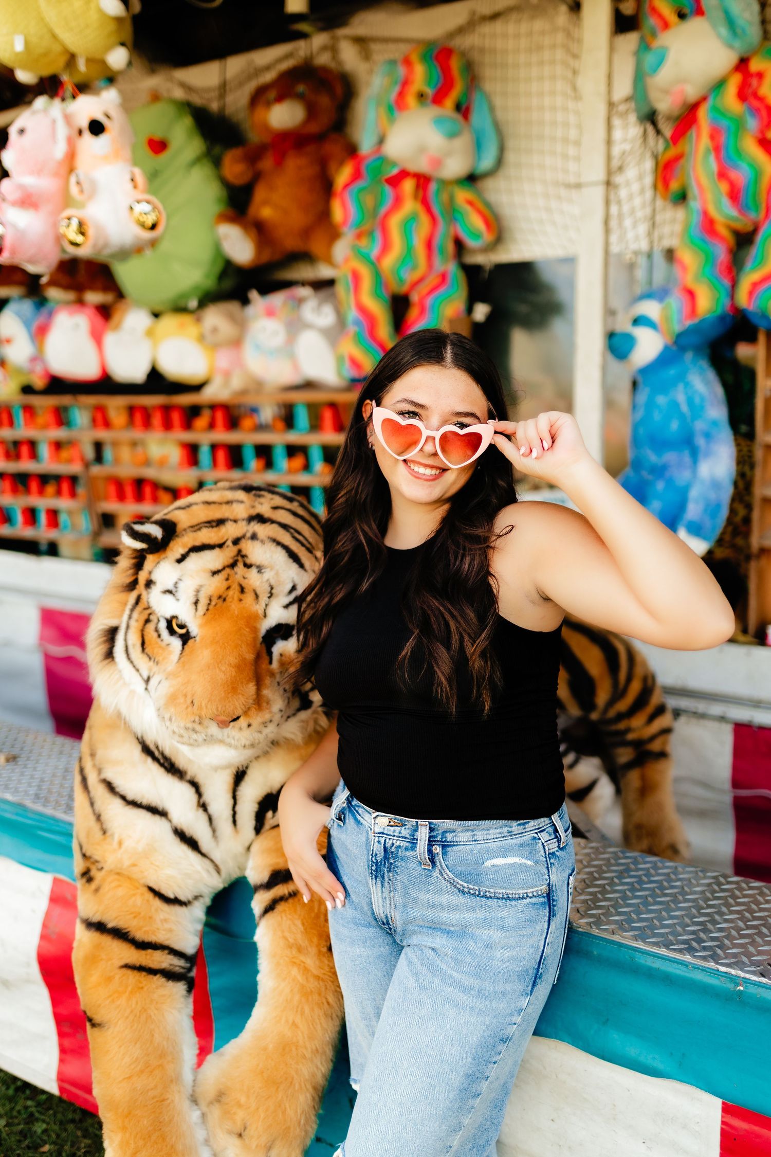 Young woman in black top with heart sunglasses posing beside a stuffed tiger at a carnival.