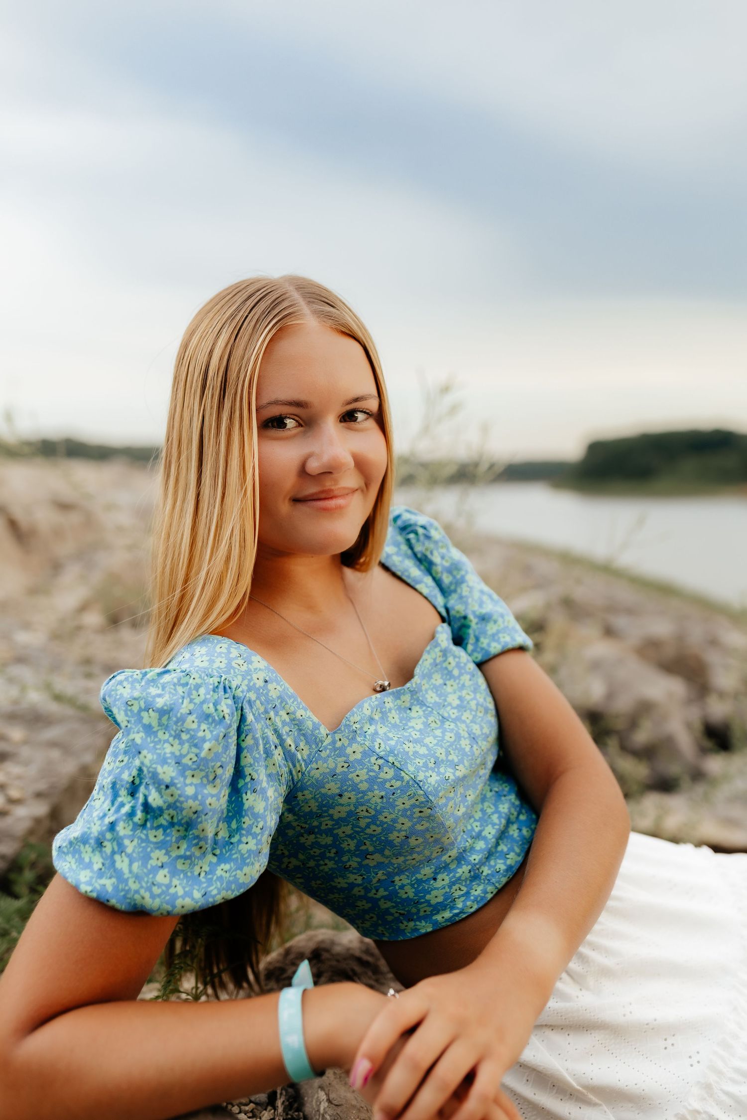 Blonde teenage girl in blue floral puff-sleeve top sits on rocks near a lake at dusk.