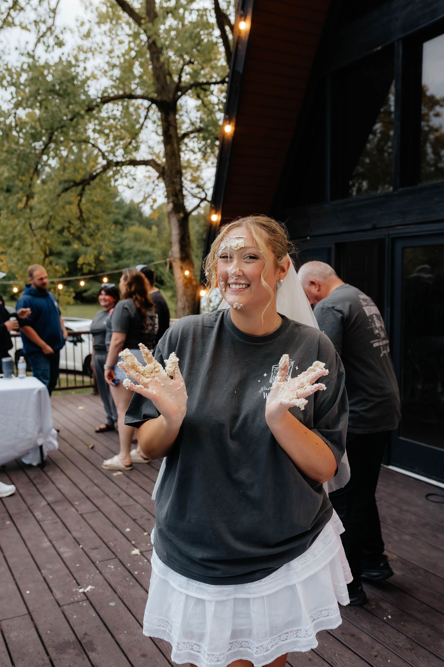 Young woman laughing with cake smeared on her face and hands at an outdoor wedding reception.