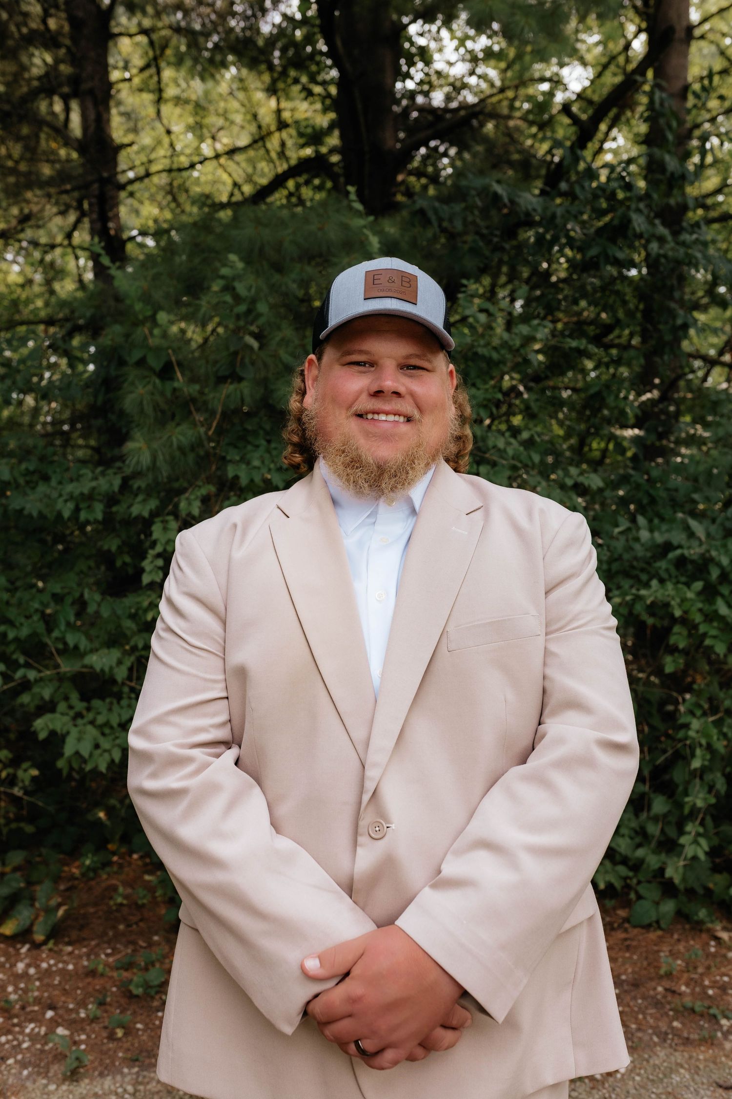 Smiling groom in beige blazer and blue leather patch cap poses in front of lush green trees.