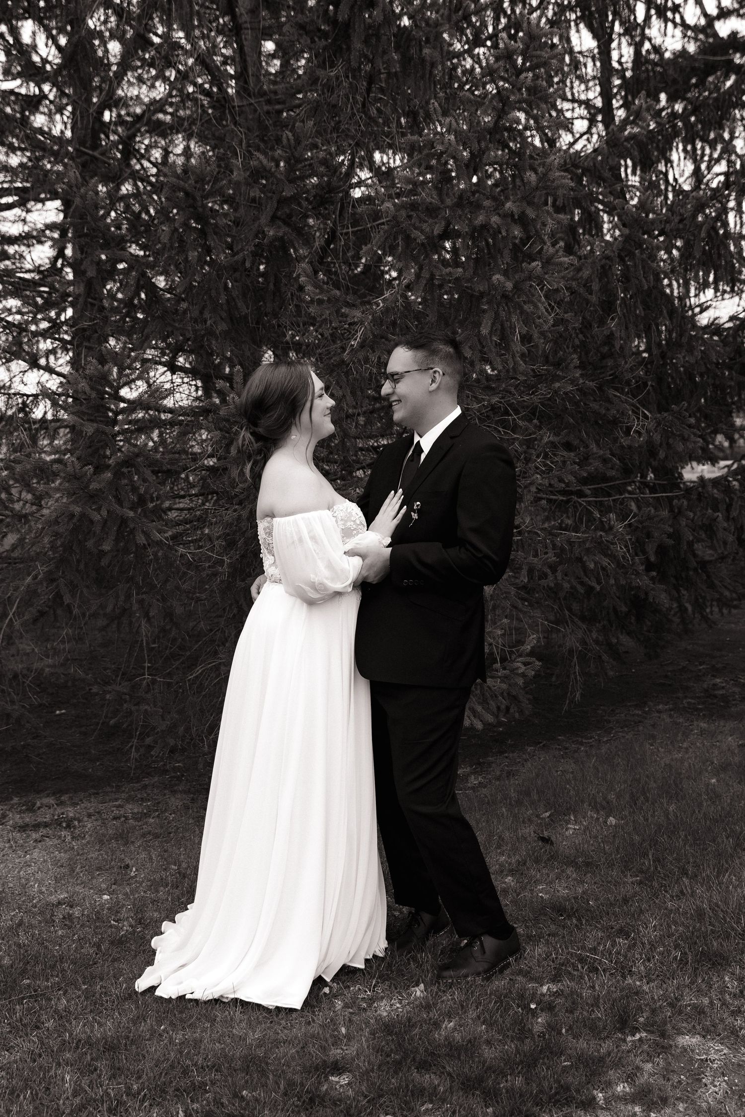 Black and white portrait of bride and groom gazing at each other lovingly beneath tall evergreen trees.