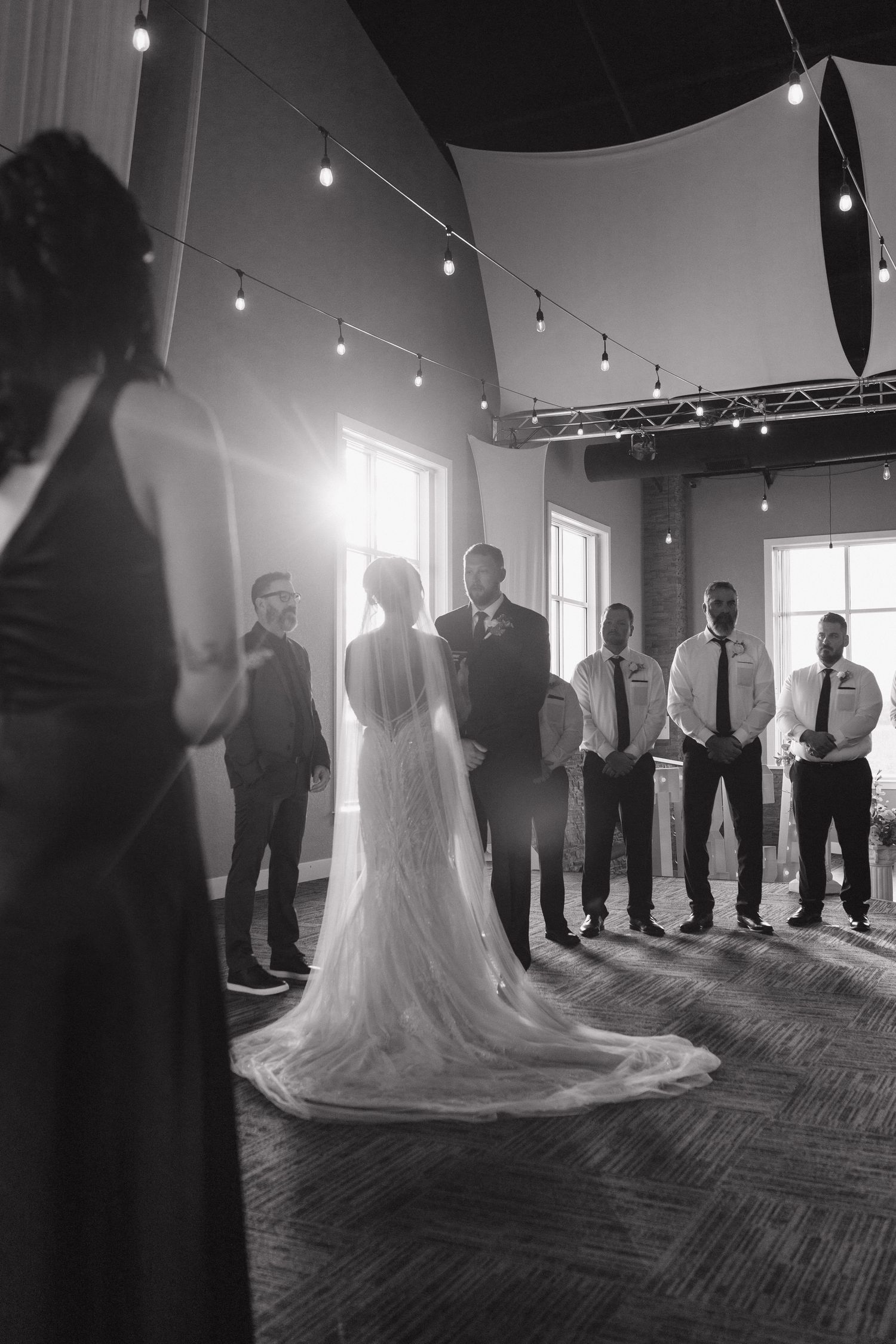 Black and white photo of a bride in a lace gown facing groomsmen during an indoor wedding ceremony.