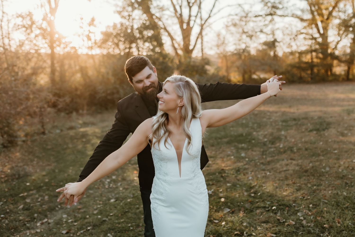 Bride in white gown and groom in black suit dancing outdoors at golden hour with arms outstretched.