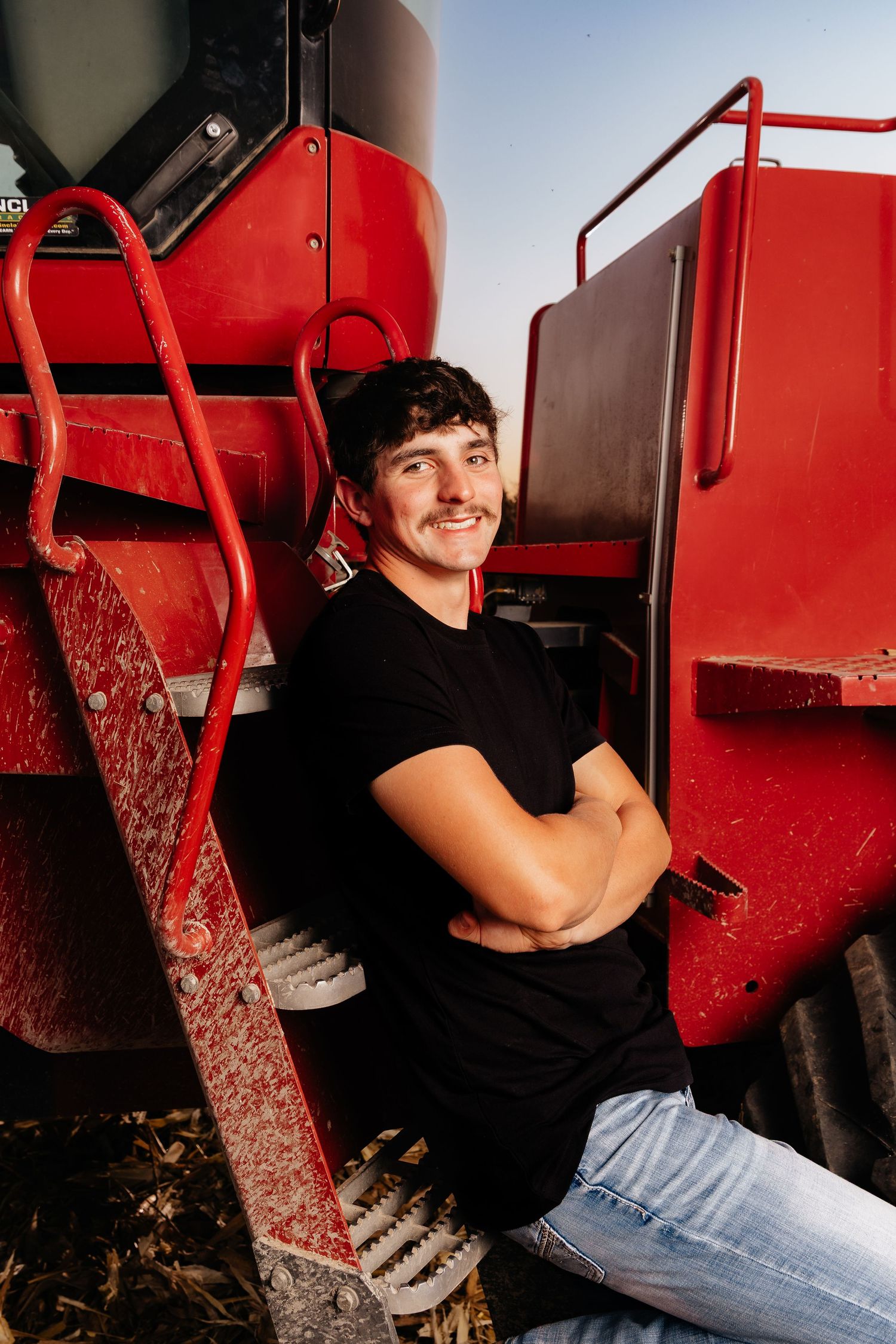 Smiling young man in black shirt sits with arms crossed on steps of red farm equipment.