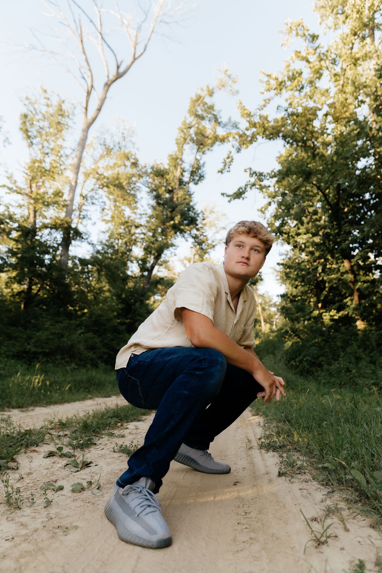 Young man in a beige shirt and jeans crouching on a sandy path surrounded by green trees.
