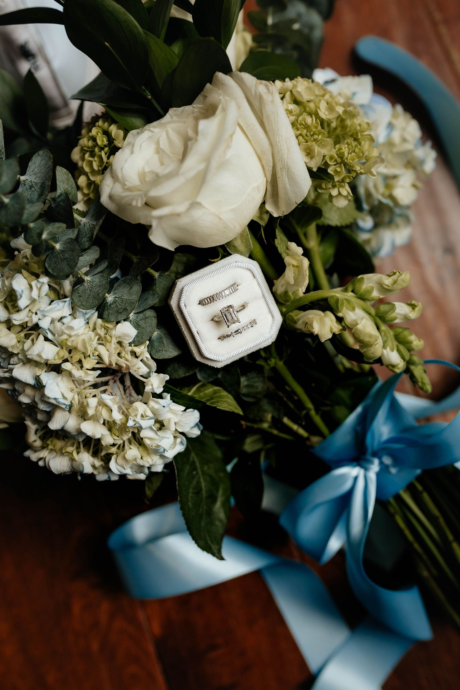 Diamond wedding ring set in white velvet box nestled in white rose and hydrangea bridal bouquet.